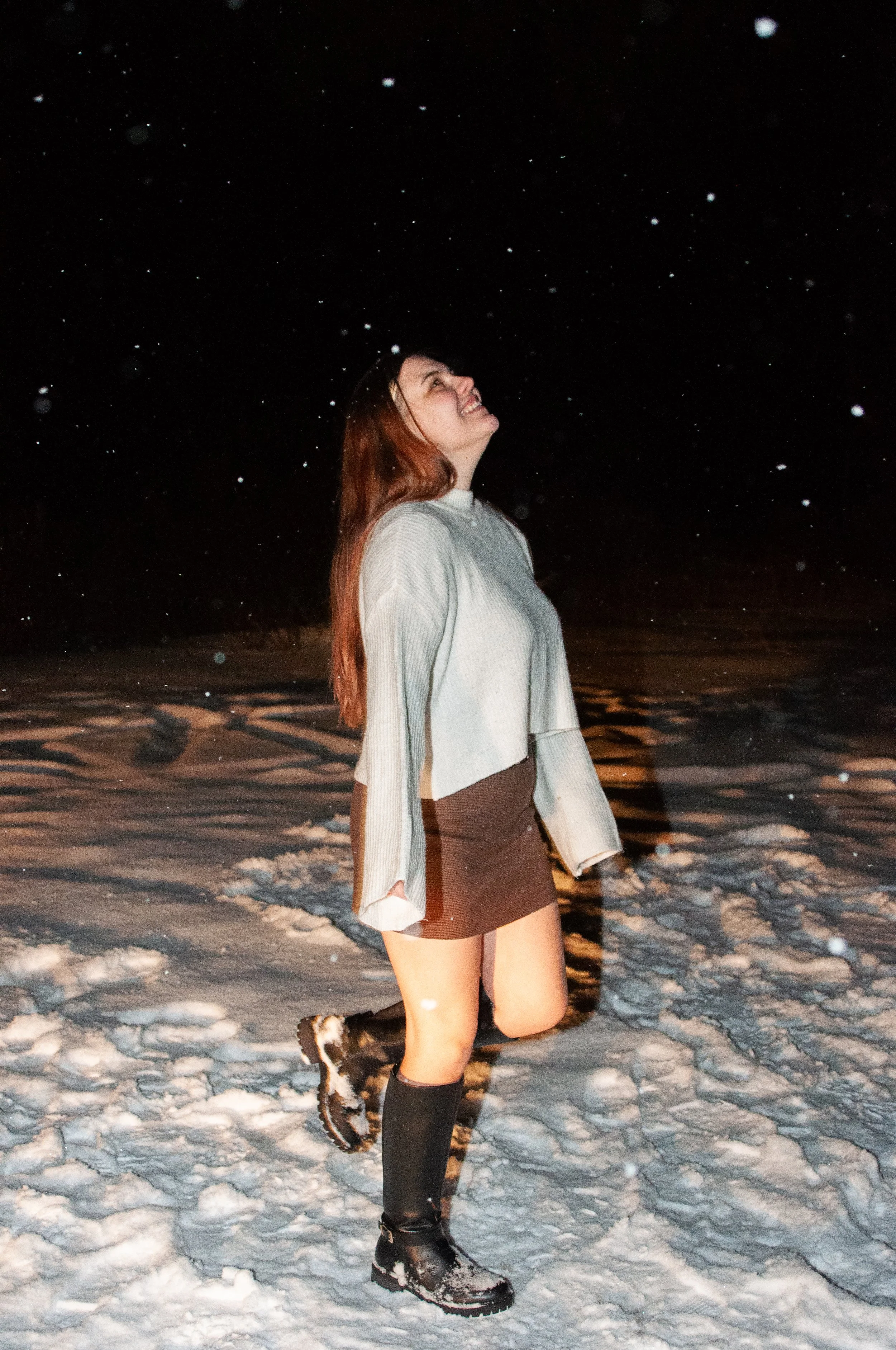 Woman in a sweater, skirt, and boots standing on snow at night, smiling and looking up at falling snowflakes.