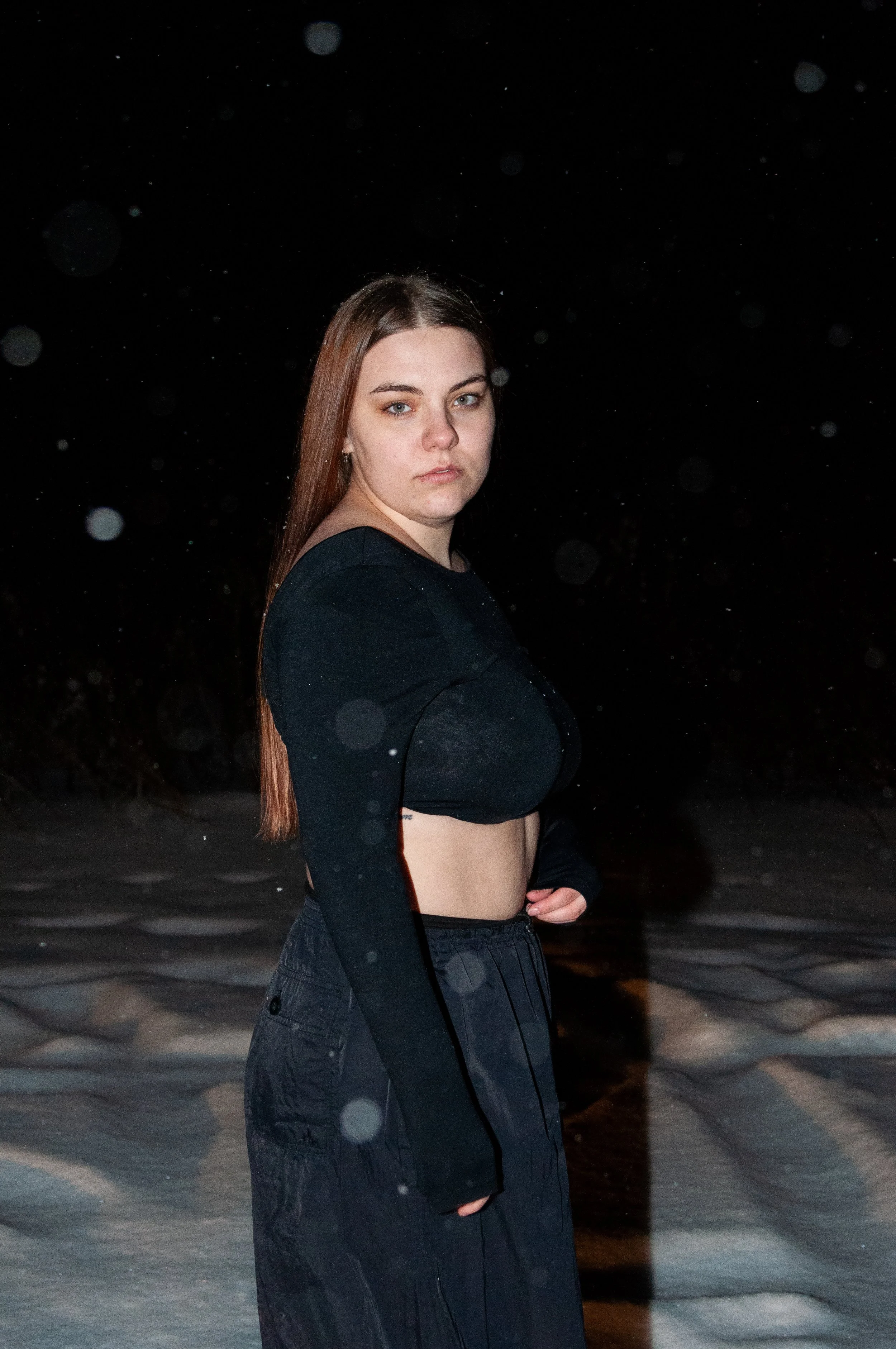A young woman standing outdoors at night in the snow, wearing a black cropped long-sleeve shirt and black pants, with snow falling around her.