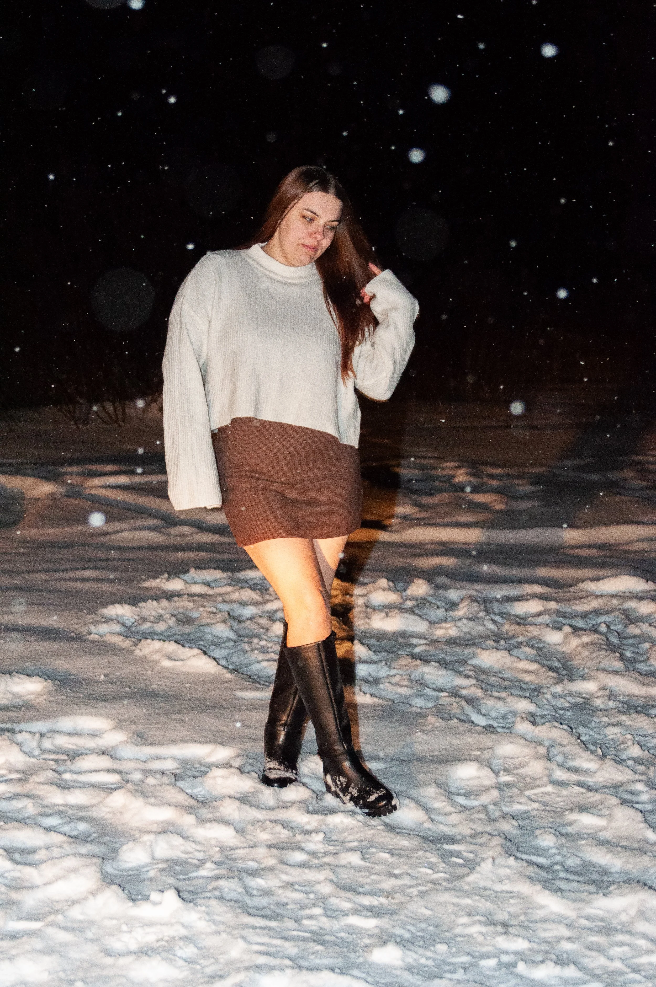 Woman standing in snow at night, wearing a white sweater, a brown skirt, and tall black boots, with snowflakes falling around her.