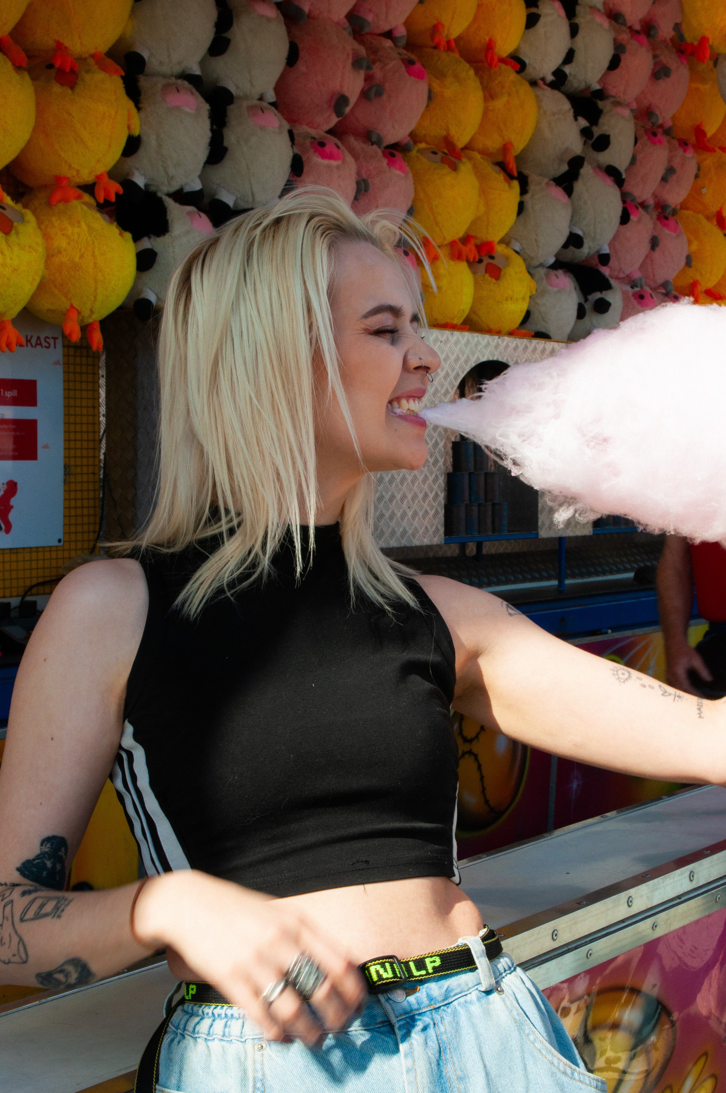 A young woman with blonde hair and tattoos is at a carnival game, exhaling a large cloud of cotton candy smoke, with a background of plush toys.