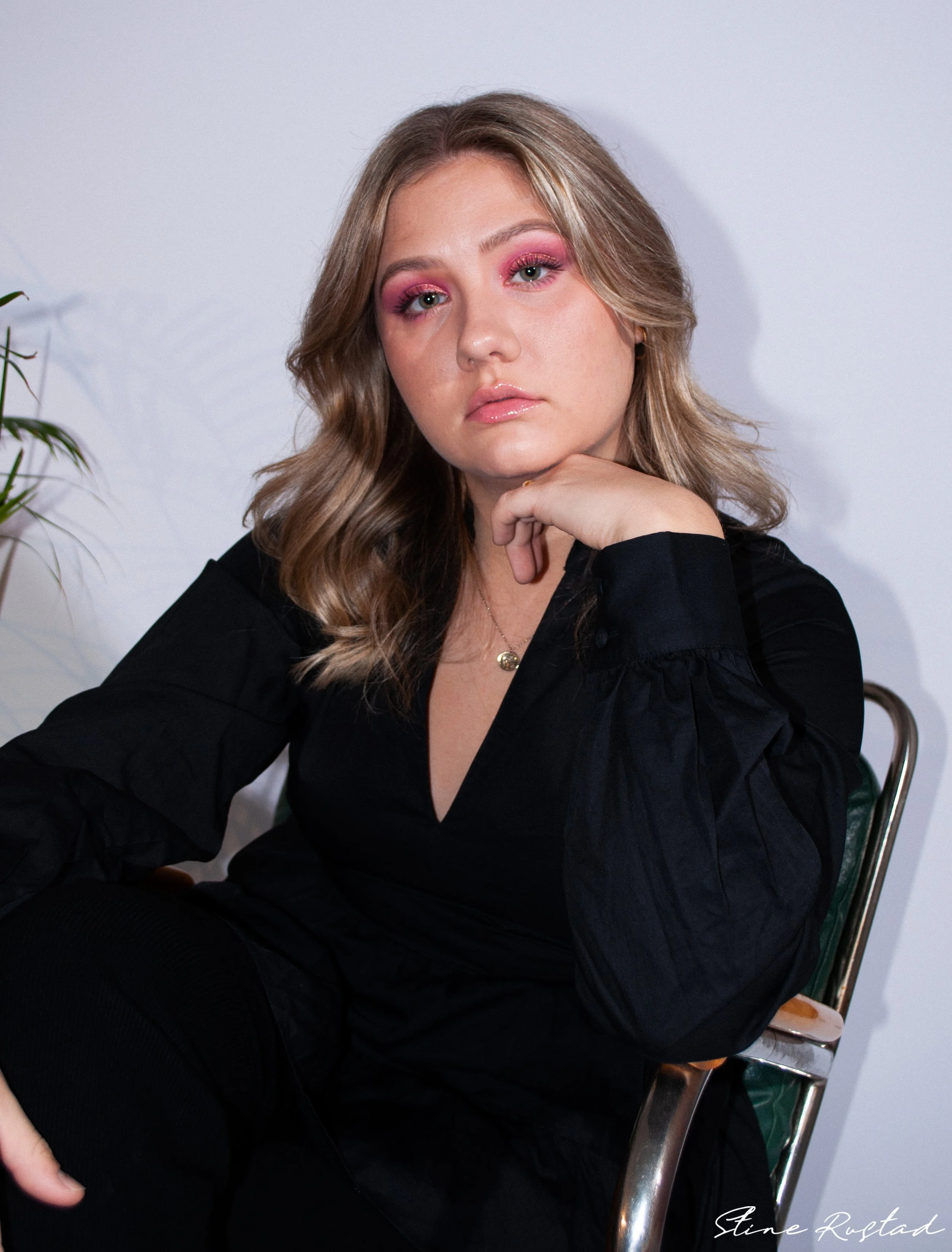 A woman with wavy blonde hair, pink eyeshadow, and glossy lips sitting on a chair against a white wall, with a plant visible on the left.