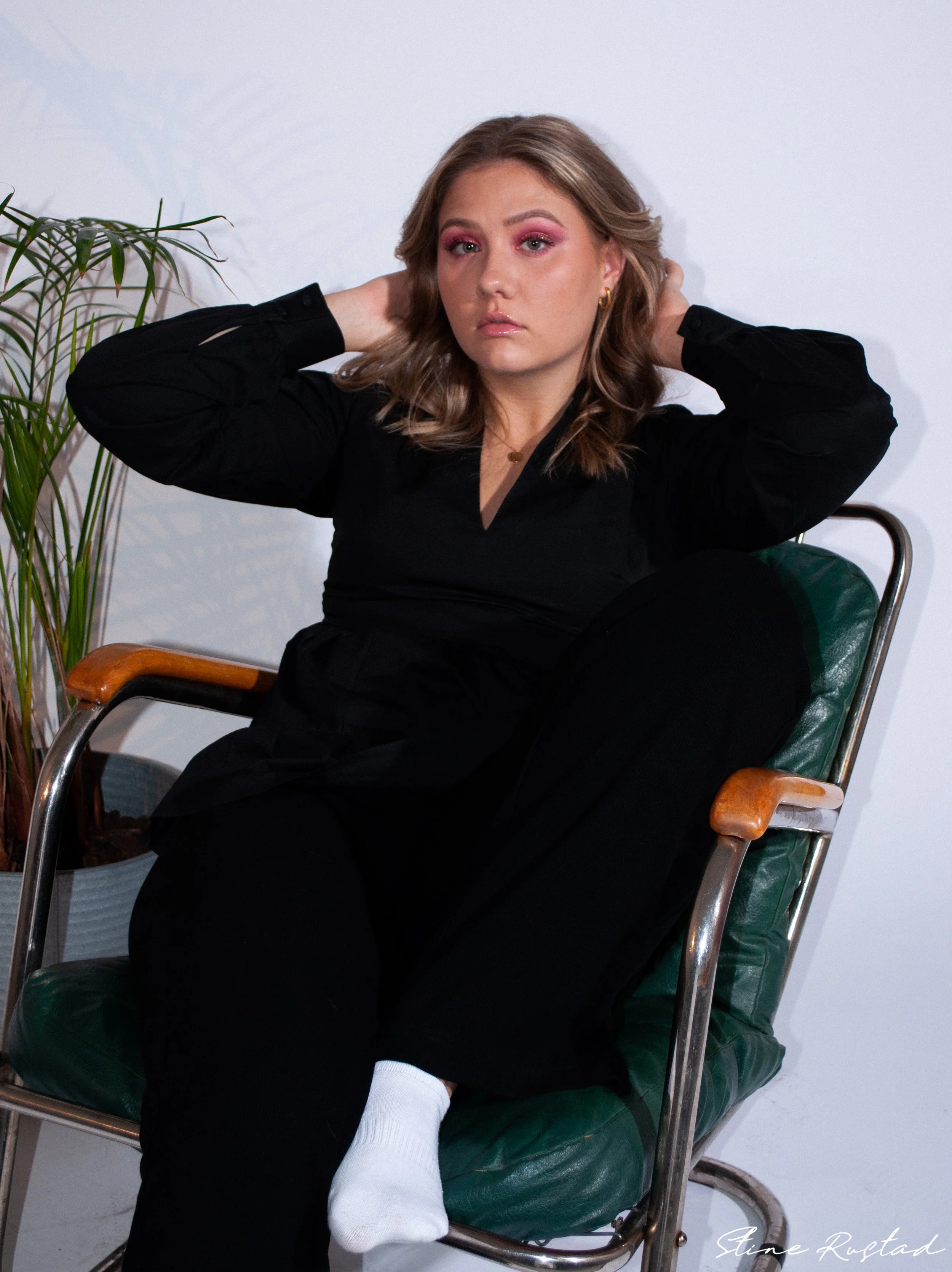 A woman with shoulder-length light brown hair, pink eyeshadow, and gold earrings, wearing a black outfit, seated in a green-cushioned chair with wooden armrests, resting her head with her hands behind her head, next to a potted plant, against a white