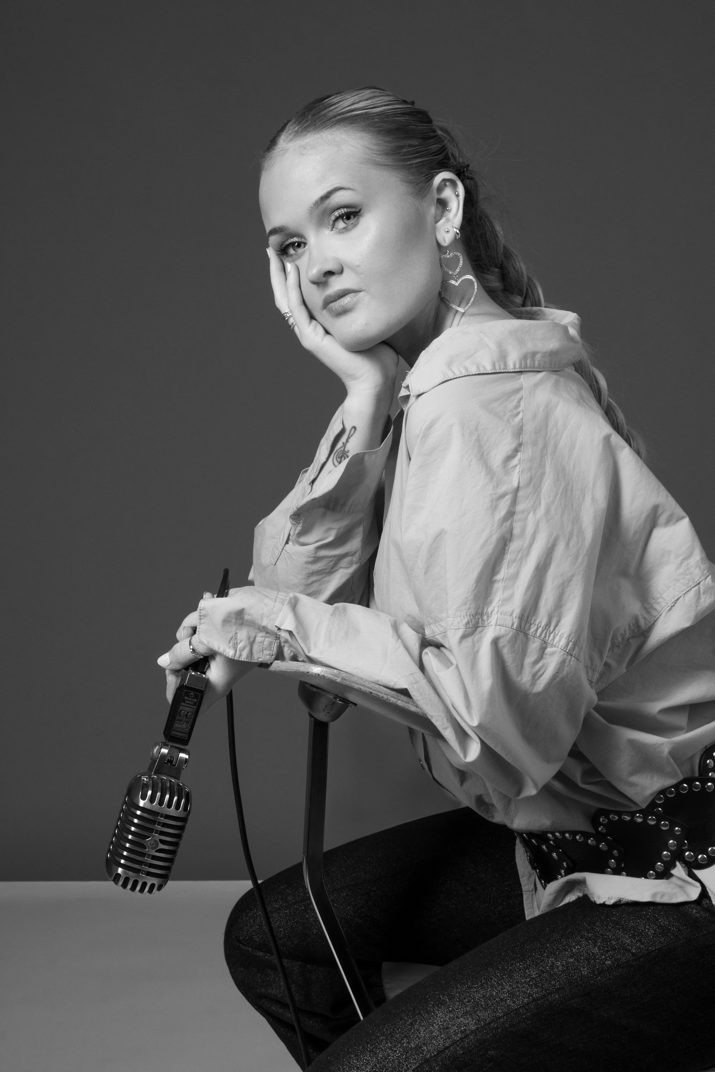 Black and white photo of a woman with braided hair, wearing earrings and a casual shirt, sitting and resting her chin on one hand while holding a vintage microphone with the other hand.