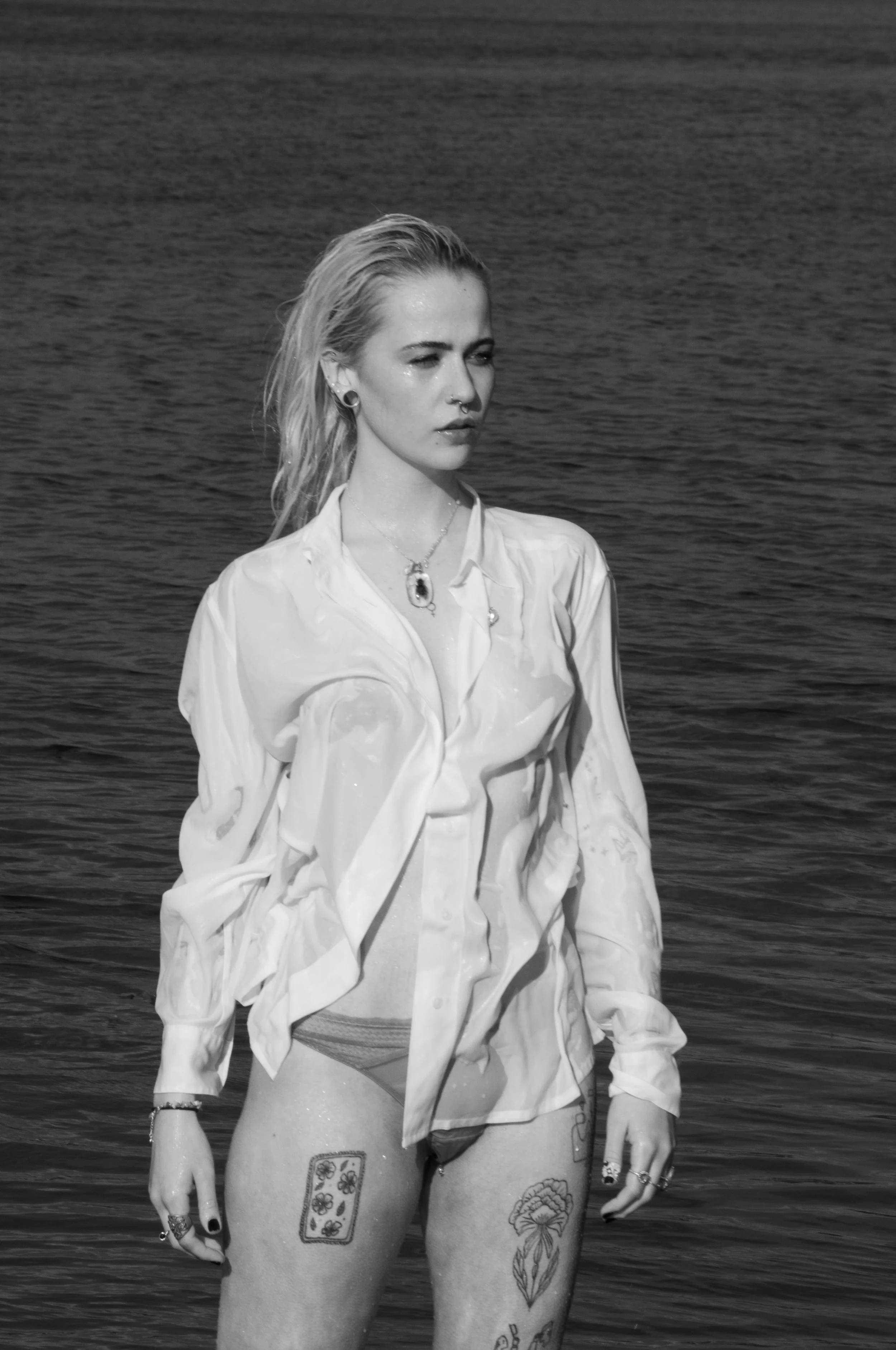 A woman with wet hair standing in the water, wearing a wet shirt and underwear, with visible tattoos on her thighs, near a large body of water, in black and white.