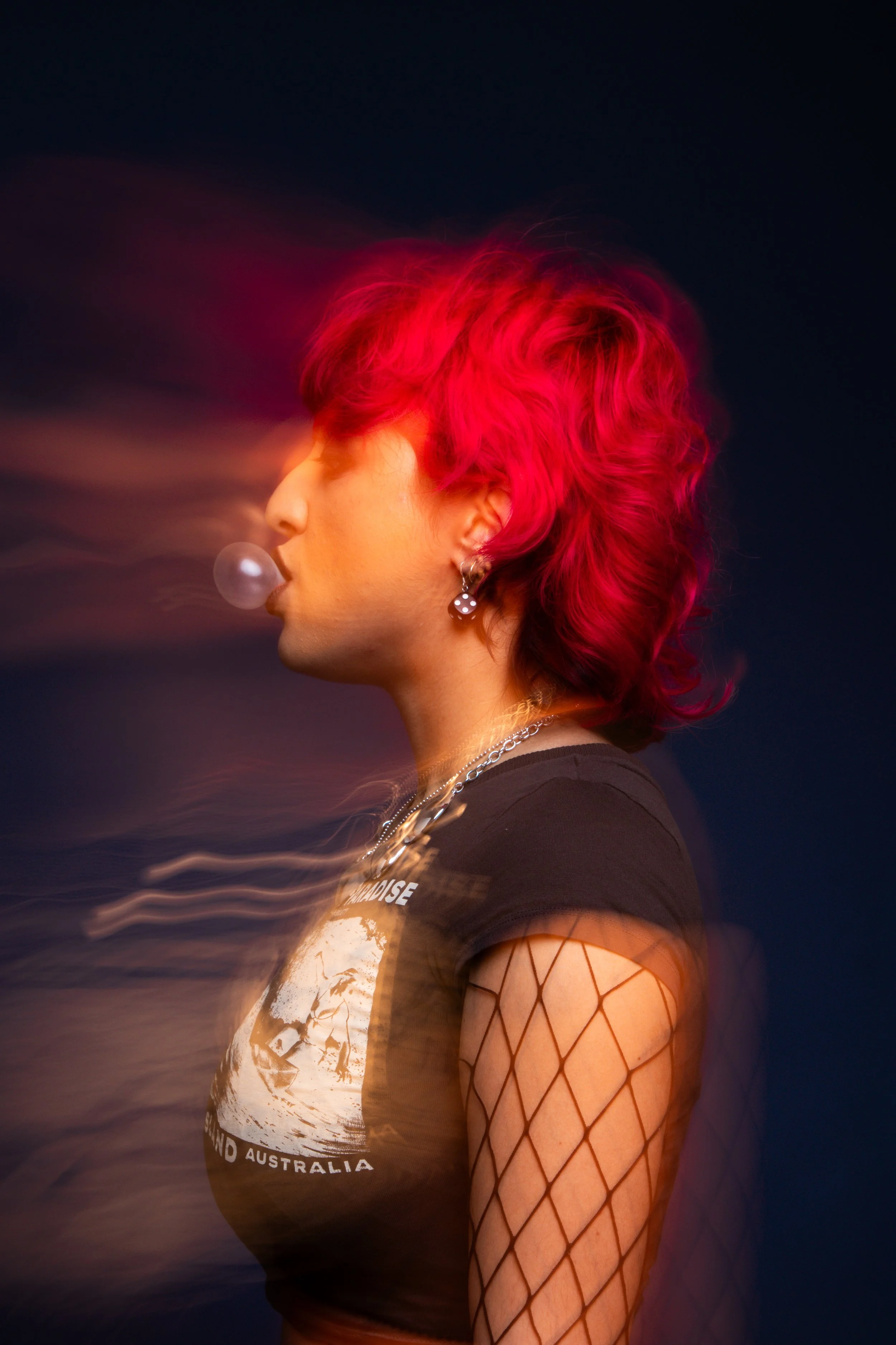 A woman with bright pink hair blowing bubble gum in profile shot, wearing a black t-shirt with a graphic of Australia, fishnet sleeves, and jewelry, set against a dark background with motion blur effects.