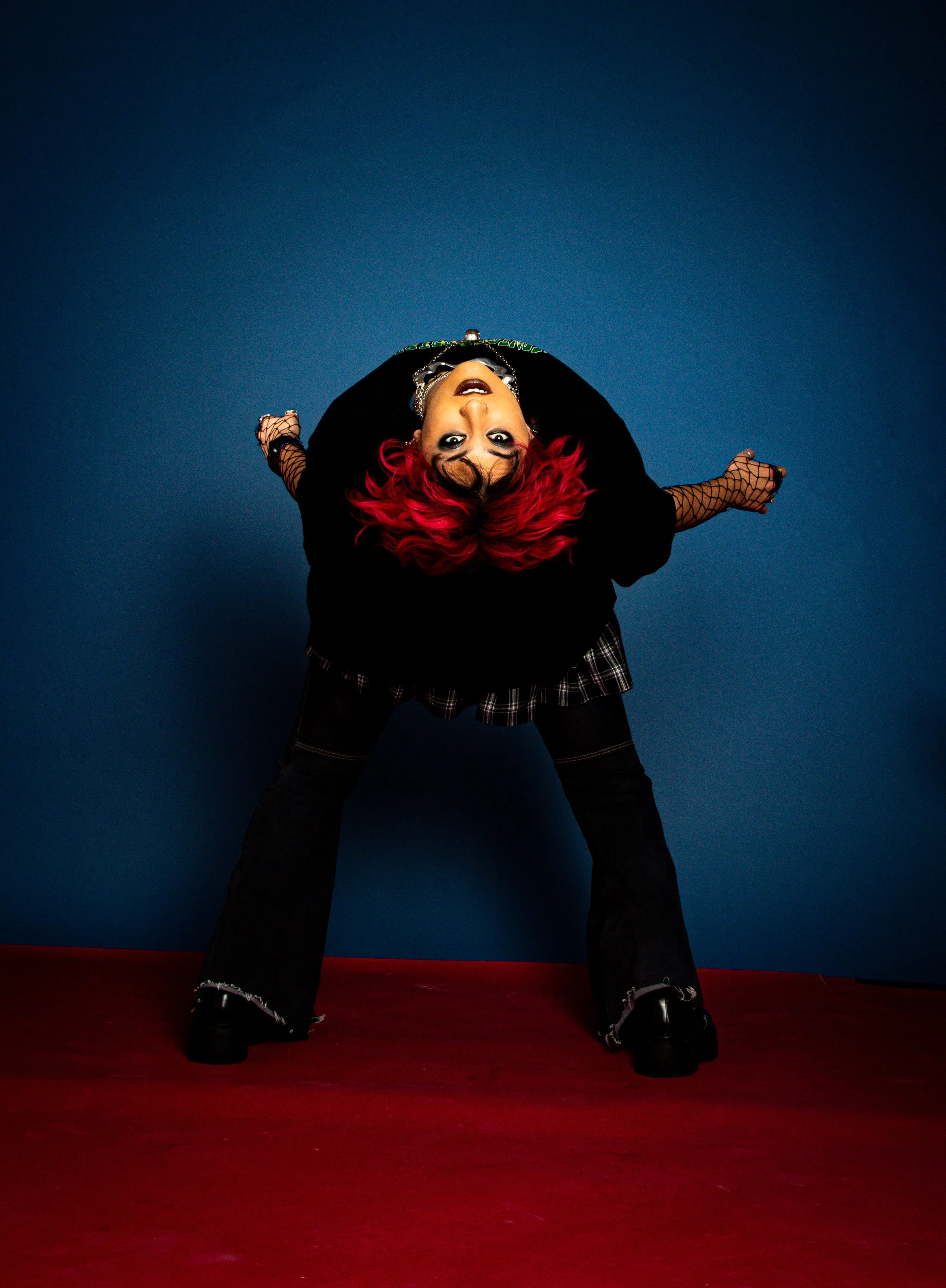 A woman with bright red hair leaning backward against a blue wall and red floor, wearing a black jacket, fishnet sleeves, plaid skirt, black pants, and platform shoes, with makeup and jewelry.