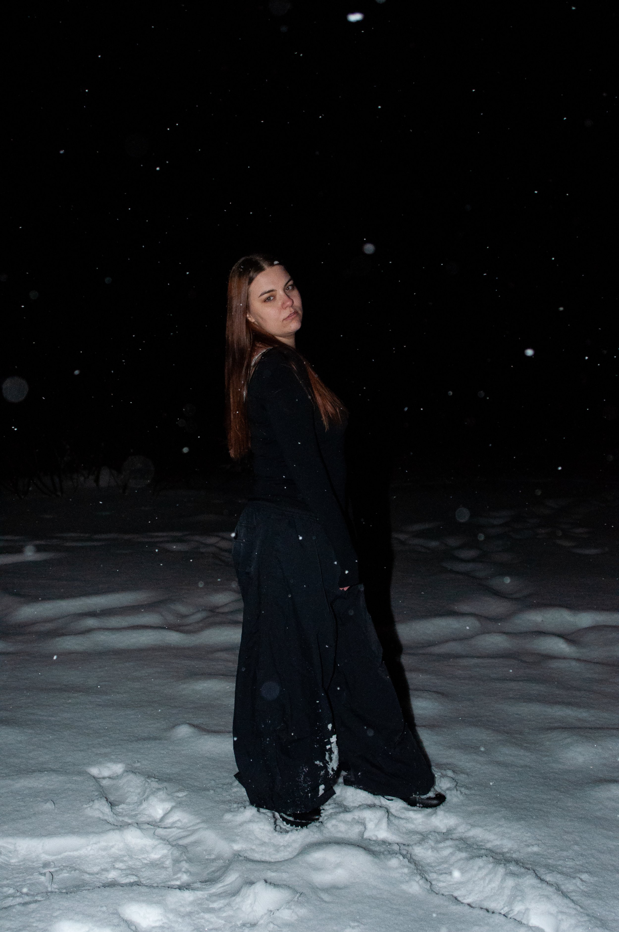 A young woman stands in the snow at night, wearing dark clothing, with snow falling around her.