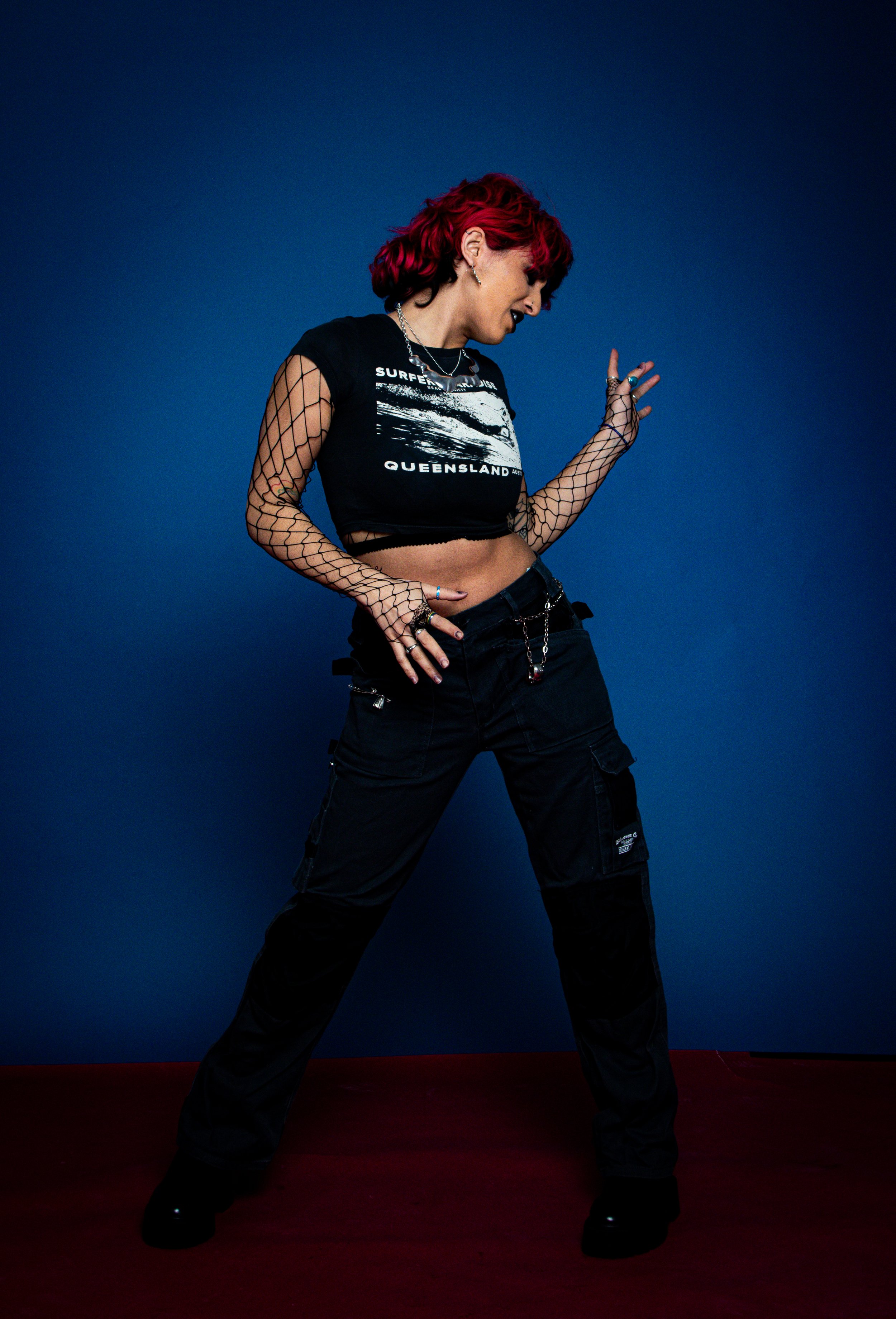 A woman with red hair in a edgy fashion pose against a blue background, wearing black cargo pants, a graphic crop top, fishnet sleeves, and various jewelry.