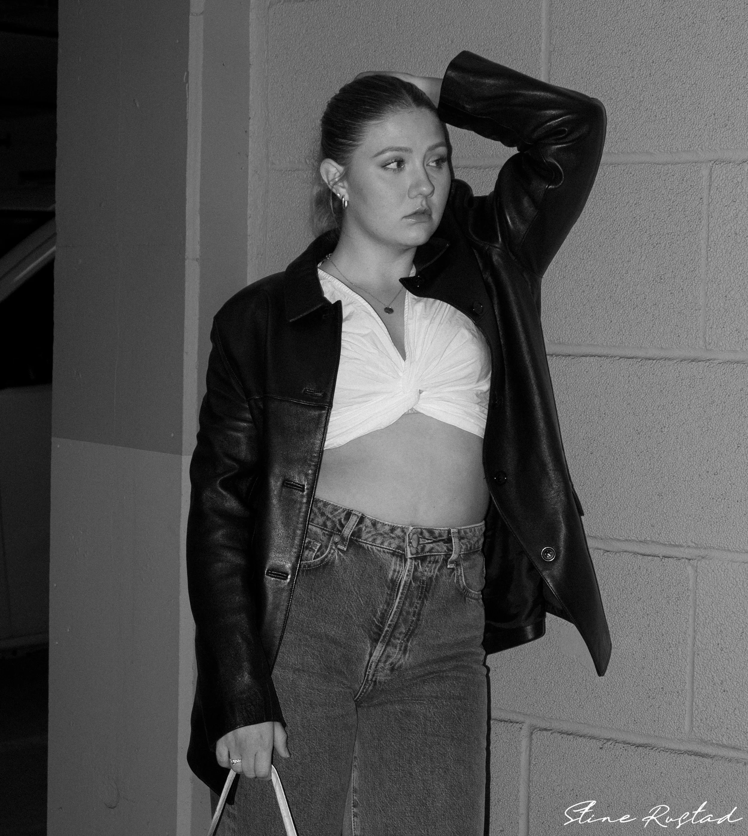 A young woman stands against a concrete wall, wearing a leather jacket over a knotted white crop top and high-waisted jeans, with her hair slicked back.