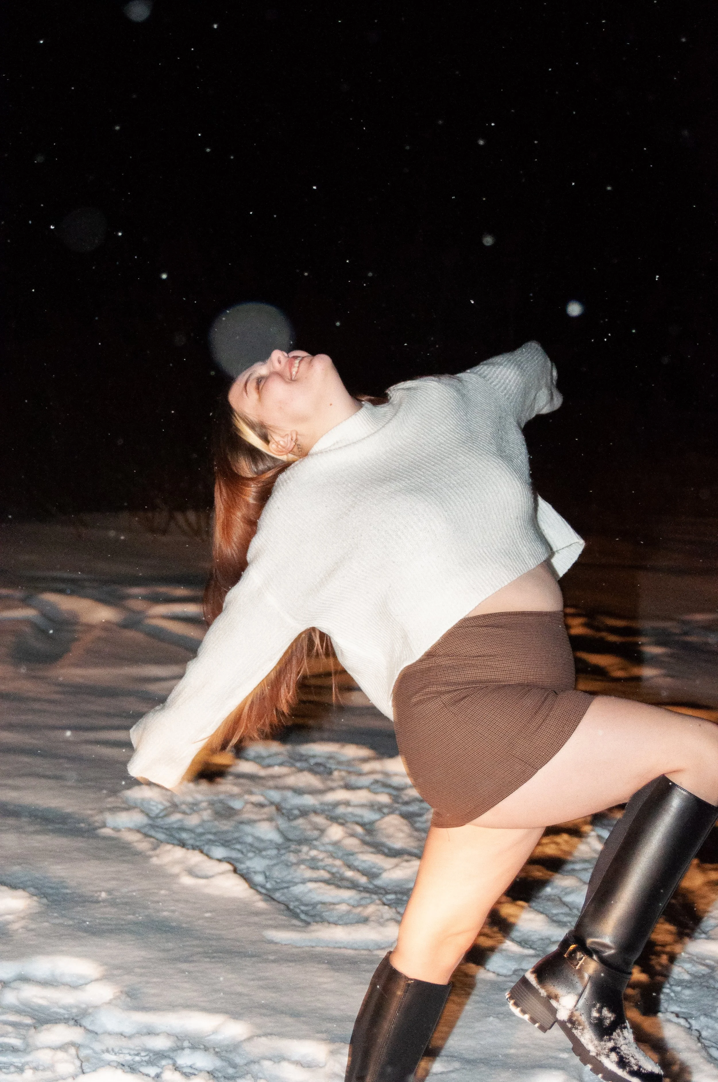 A woman smiling and leaning back on snow at night, wearing a gray sweater, brown skirt, and black boots.
