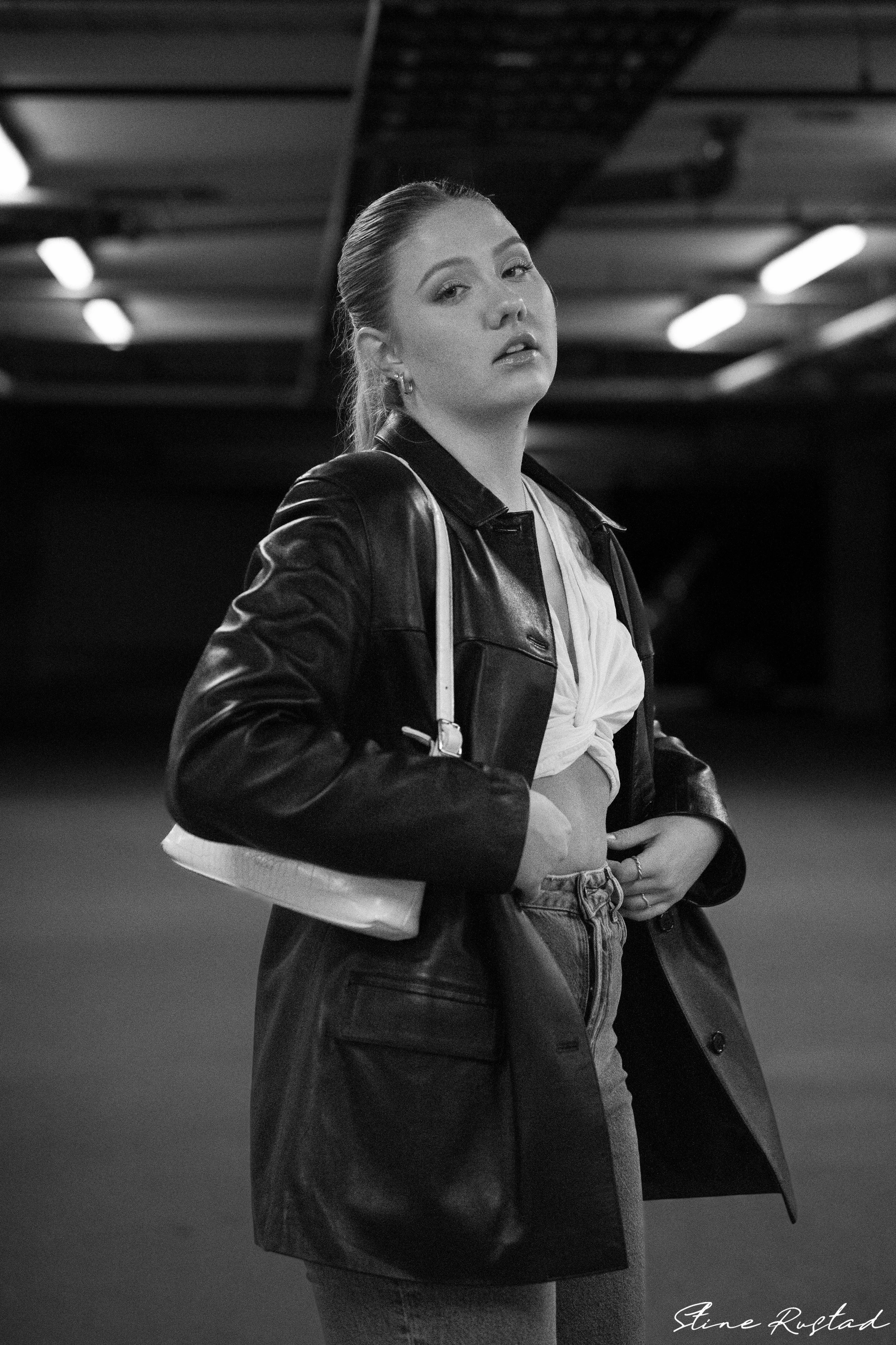 A young woman with slicked-back hair wearing a leather jacket and jeans, standing in a parking garage with overhead lighting.