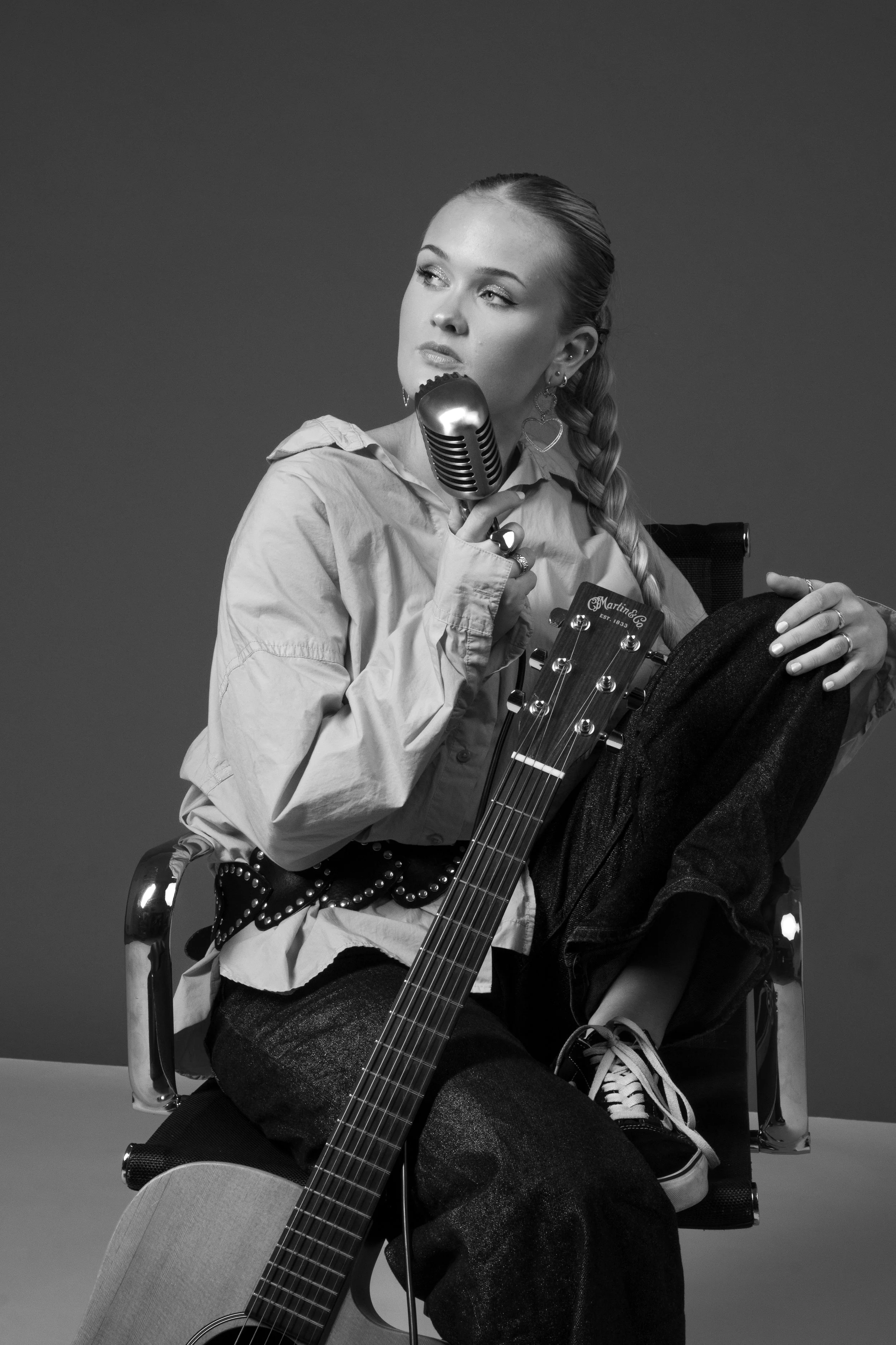 Black and white photo of a young woman sitting on a chair, holding an old-fashioned microphone near her face. She has a braid over her shoulder, and a guitar is resting against her leg. She is wearing casual clothing, including a loose jacket and sne