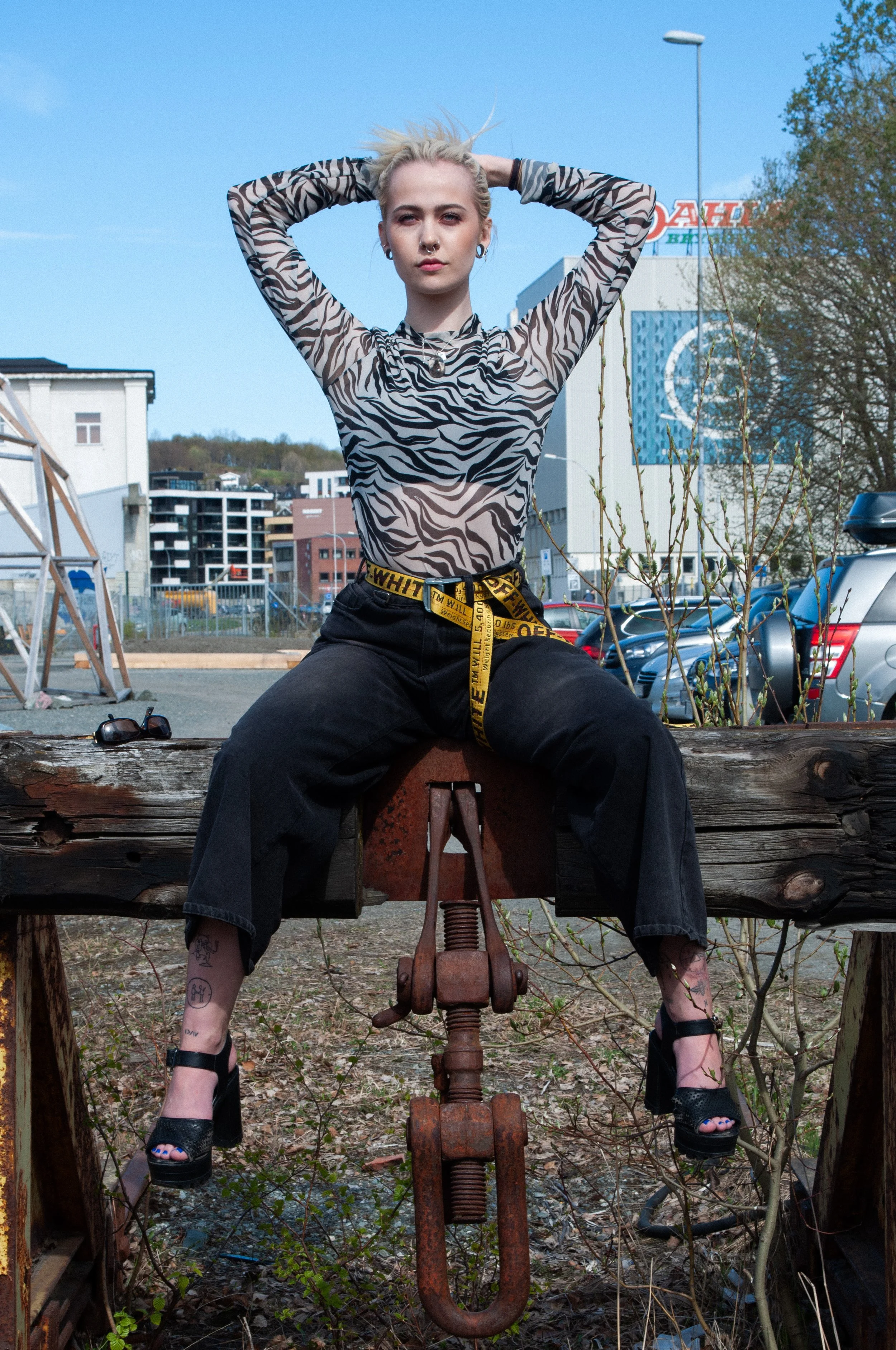 A young woman with platinum blonde hair posing outdoors, sitting on a rusted metal contraption, wearing a zebra-patterned top, black pants, high heels, and a yellow Off-White belt, with sunglasses on the ground nearby.