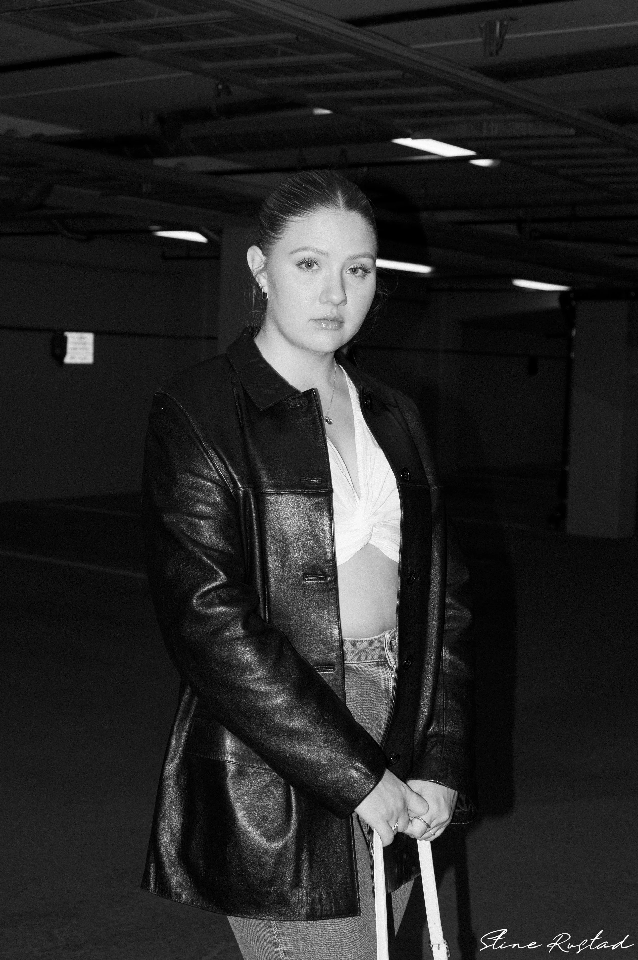 A young woman with orange hair tied back standing in an underground parking garage, wearing a black leather jacket over a white knotted top, with her hands clasped in front of her.