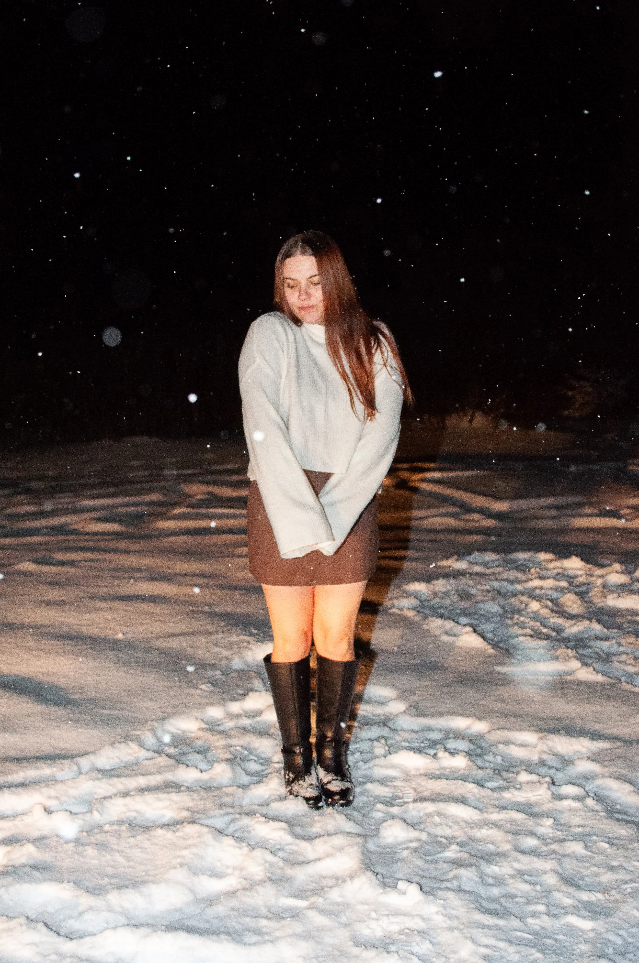 A young woman standing in the snow at night, wearing a beige sweater, a dark skirt, and tall black boots, with snow falling around her.