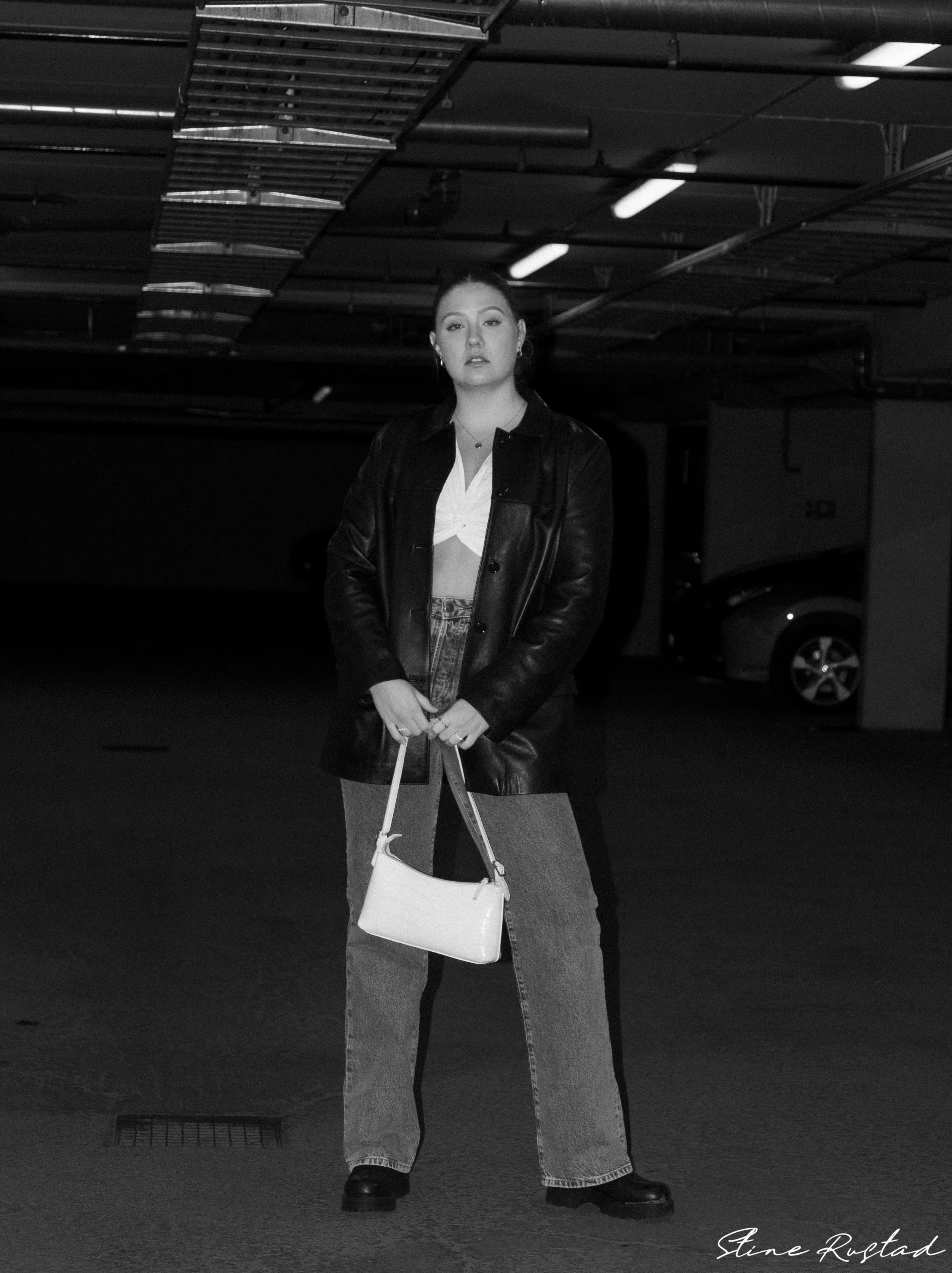 A young woman standing in a parking garage, holding a white purse, wearing a leather jacket, crop top, and jeans.