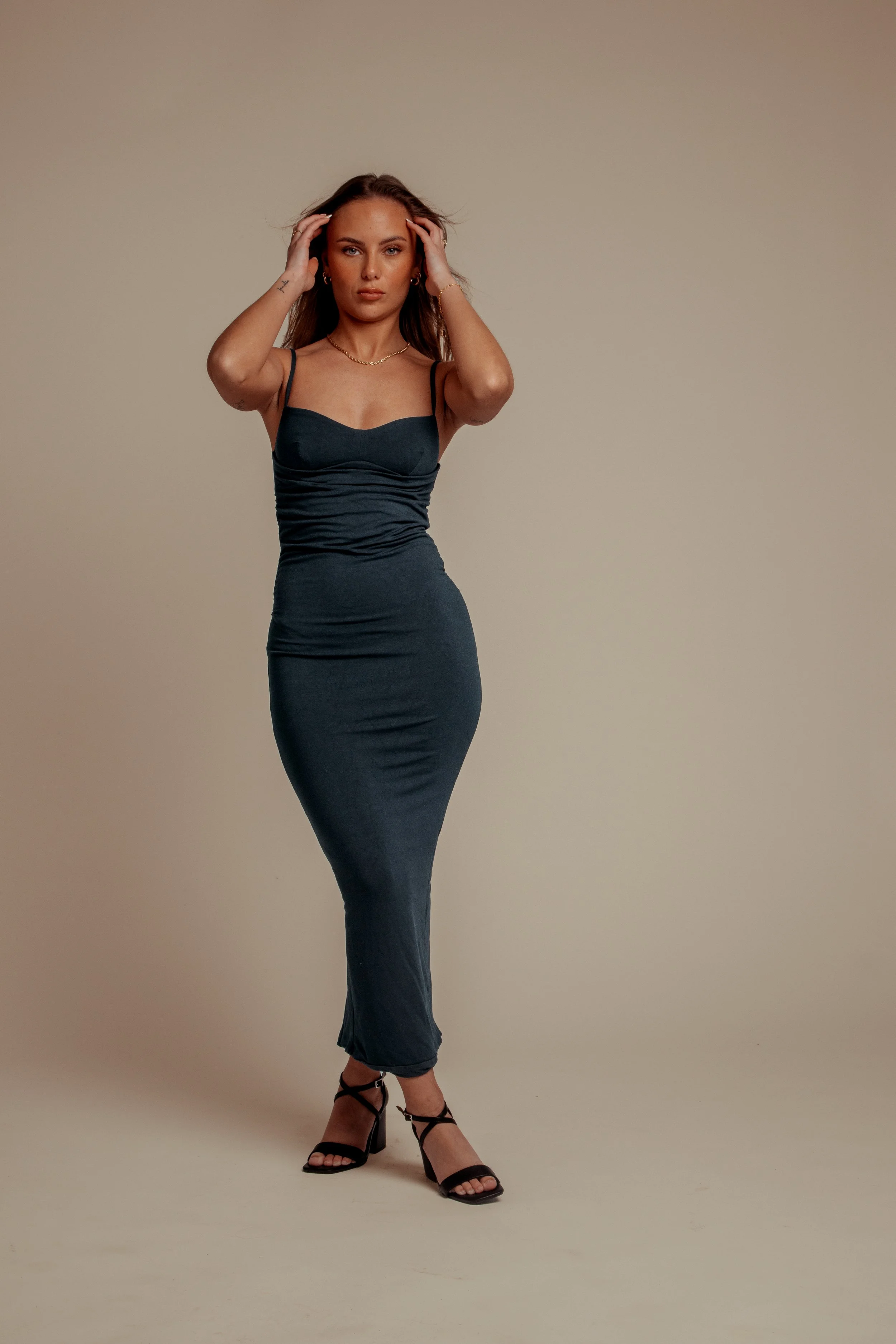 A woman with long brown hair wearing a dark gray, sleeveless, fitted dress and black high-heeled sandals standing against a plain beige background.