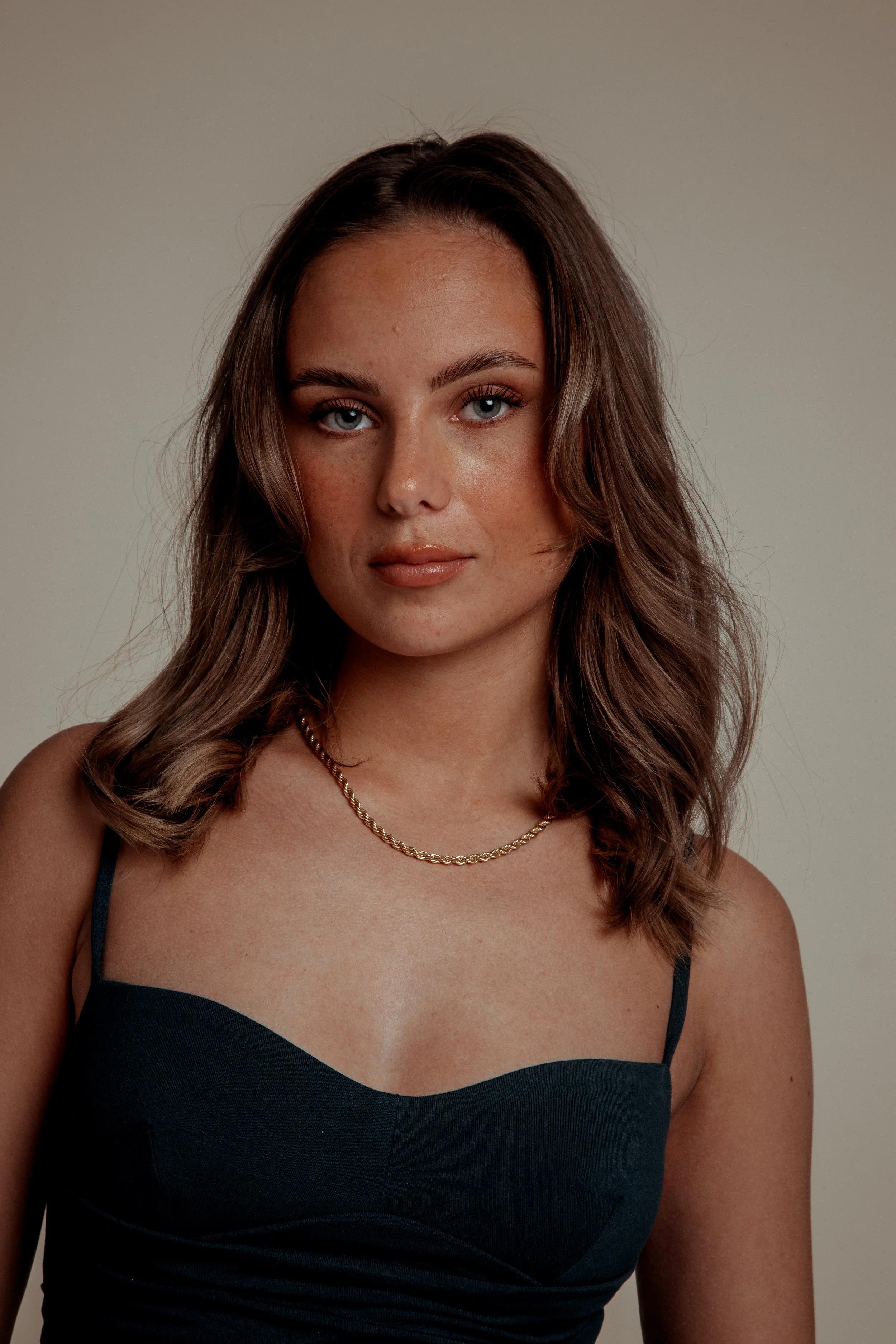 A young woman with shoulder-length wavy brown hair and blue eyes, wearing a black spaghetti strap top and a gold chain necklace, looking directly at the camera against a neutral background.