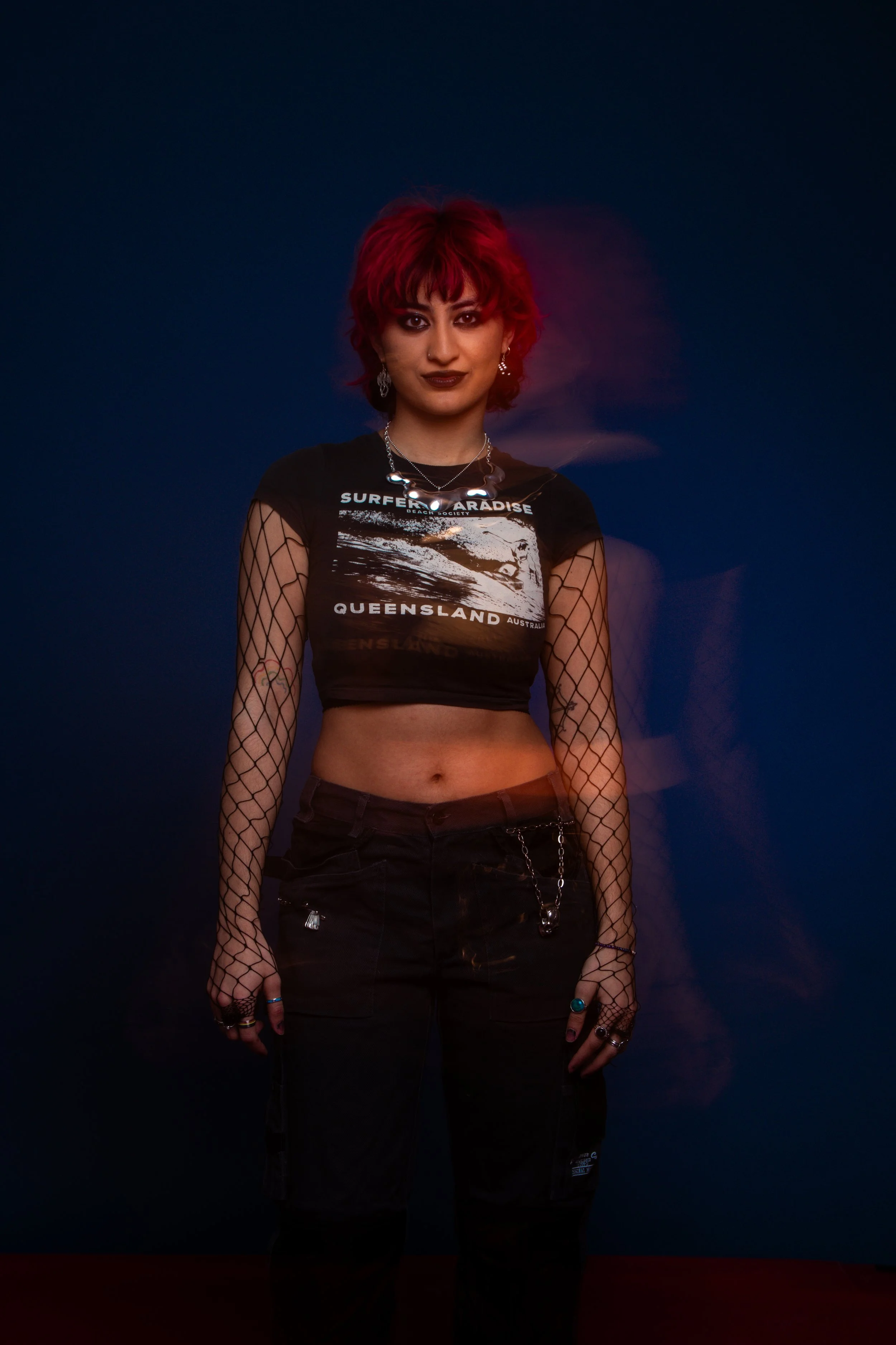 A young woman with red hair, wearing a black graphic crop top, fishnet sleeves, black pants with chains, and jewelry, stands against a blue background.
