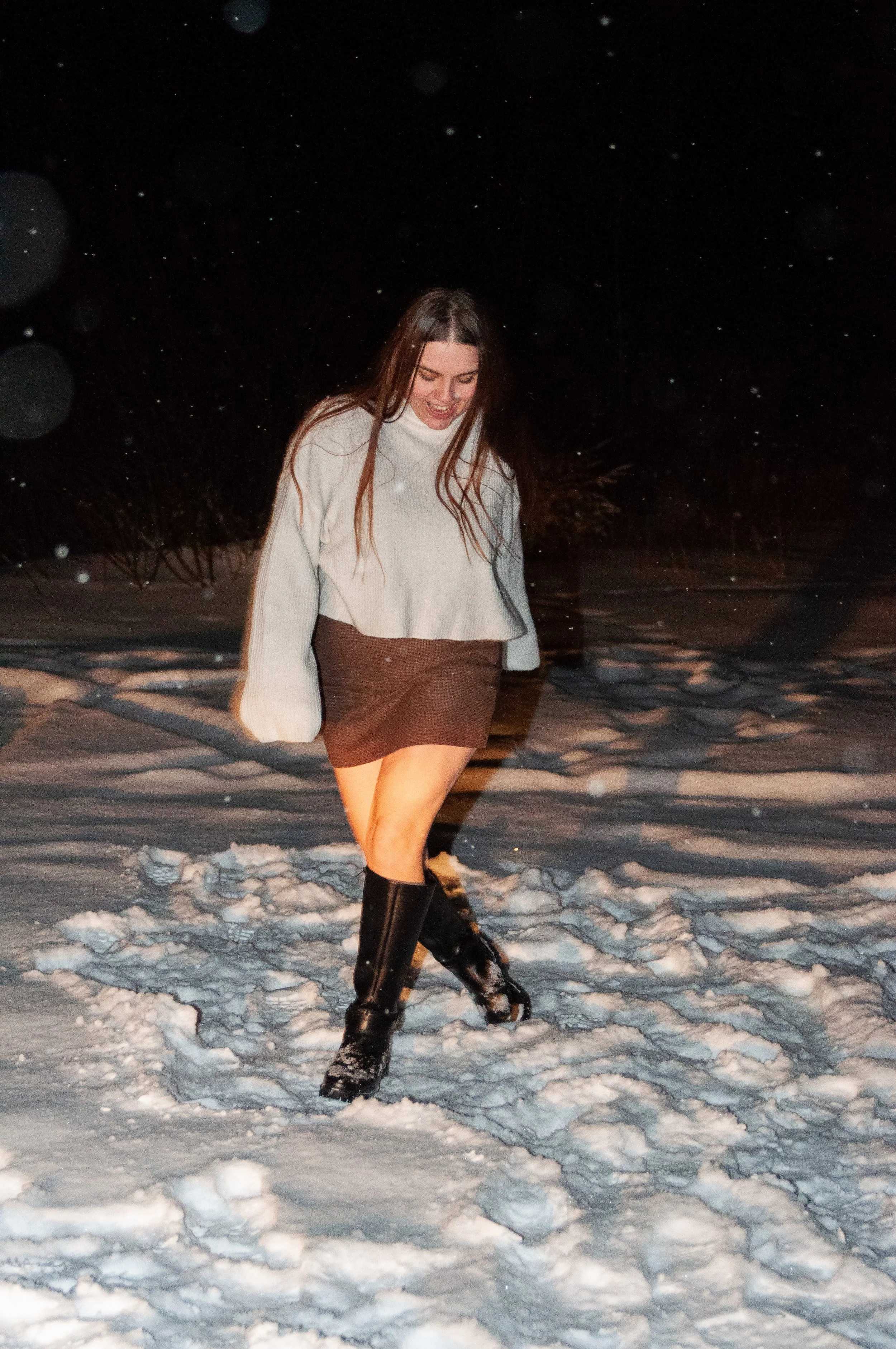 A young woman walking on snow at night, smiling, dressed in a white sweater, brown skirt, and tall black boots.