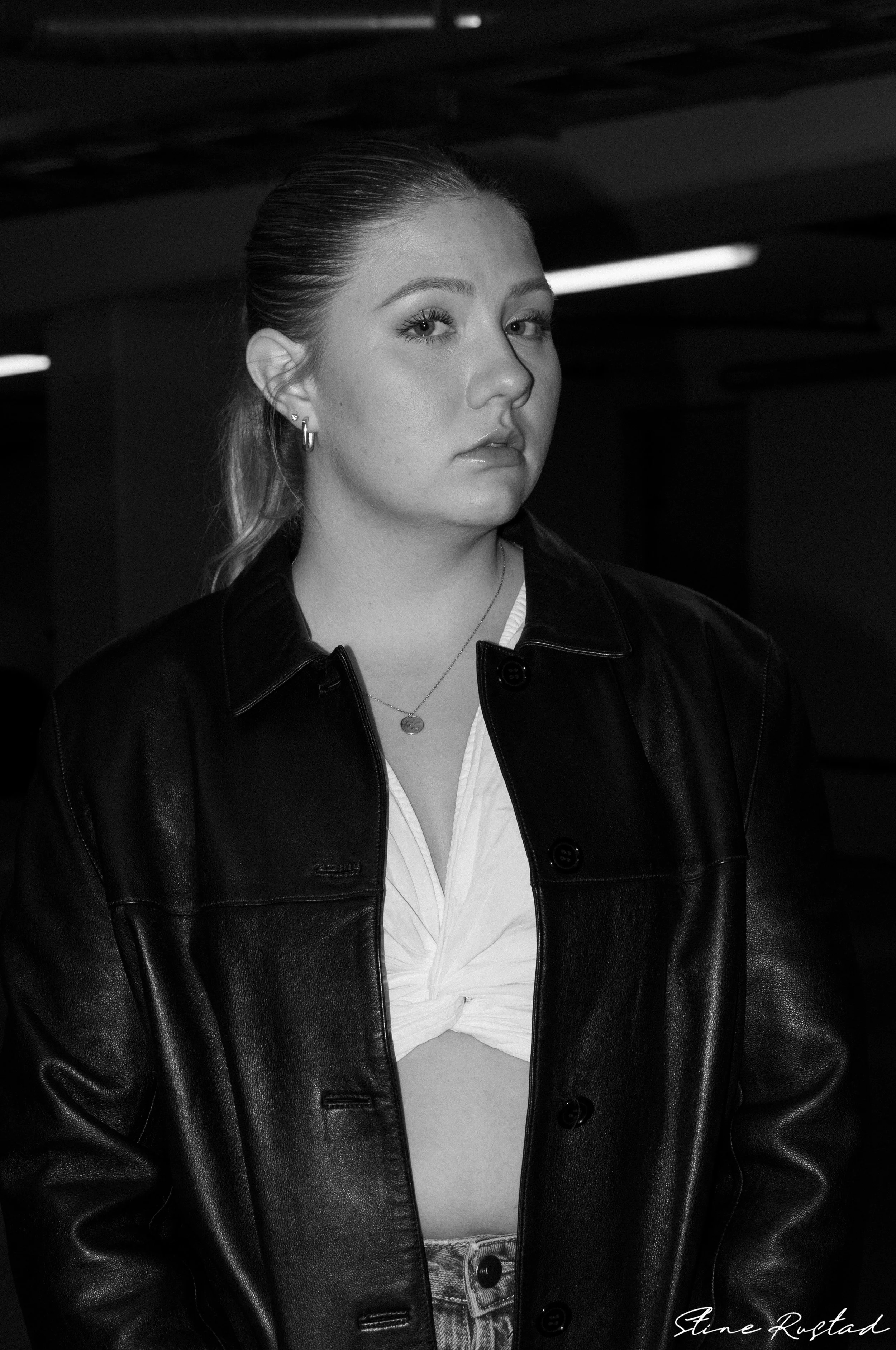 Black-and-white photo of a young woman with light skin, slicked-back hair, wearing hoop earrings, a necklace, a white knotted shirt, and a black leather jacket.