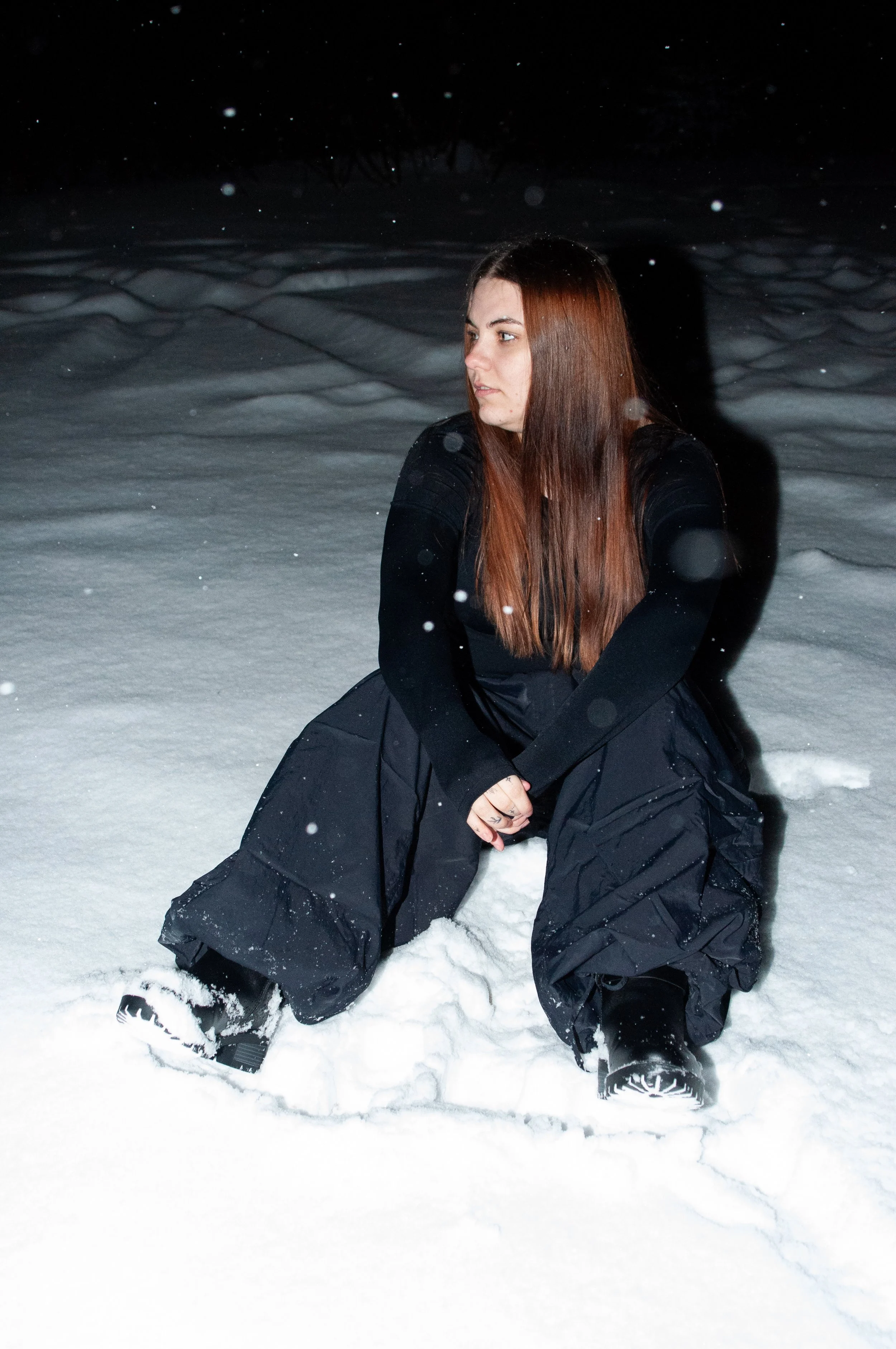 A woman with long reddish-brown hair sitting in the snow at night, wearing a black jacket and pants, with snow falling around her.