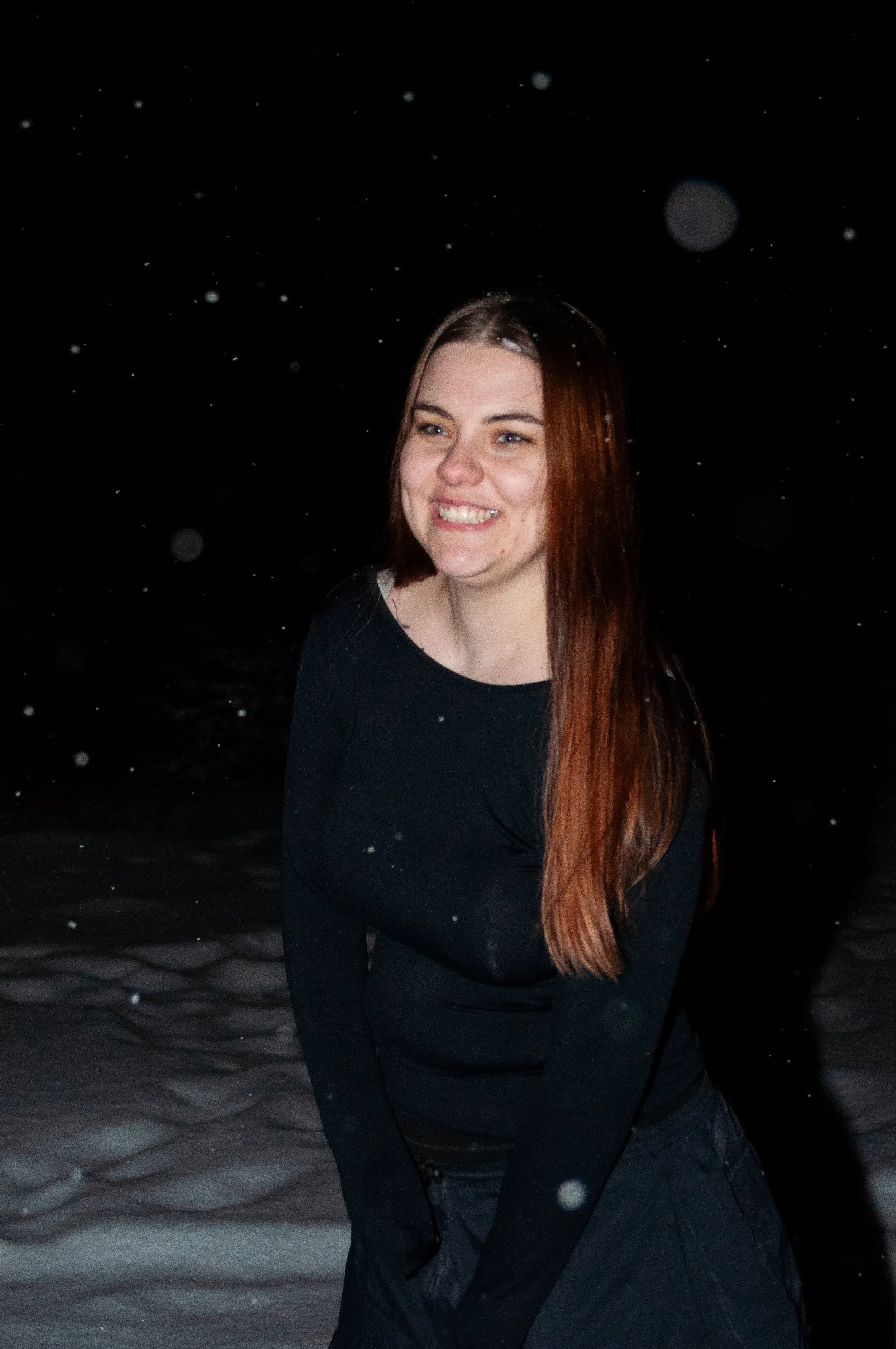 A woman with long reddish-brown hair smiling at night on snowy ground with snow falling around her.