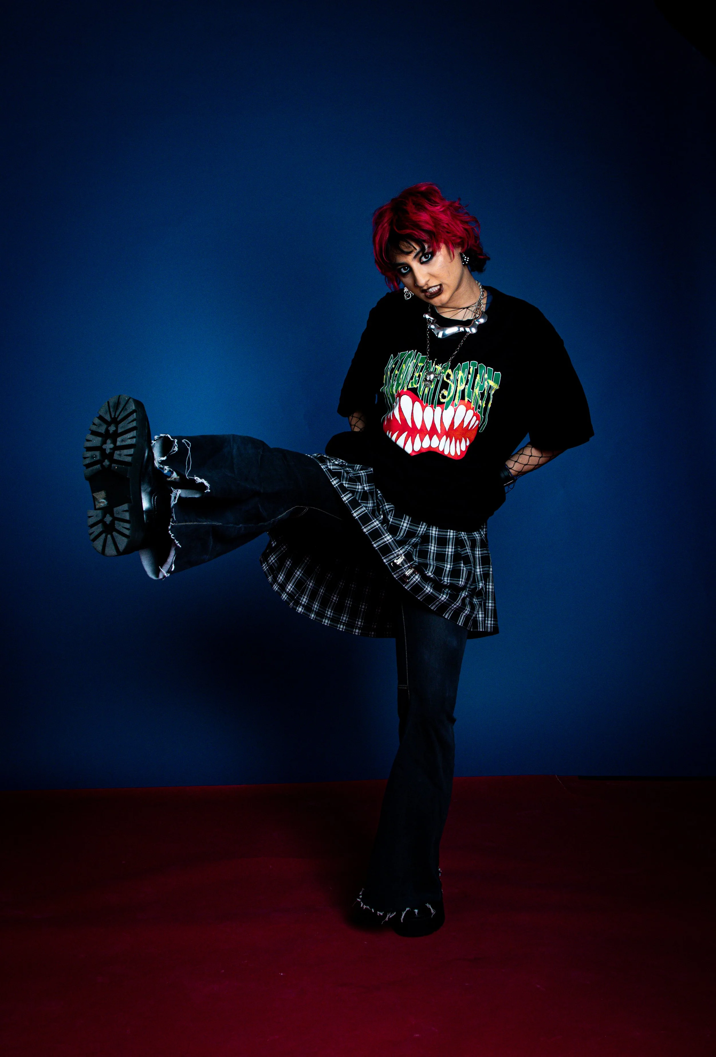 Teenager with bright pink hair, black makeup, wearing a black T-shirt with a graphic of a mouth with teeth, black ripped pants, plaid skirt, platform shoes, standing on one leg, against a blue background.