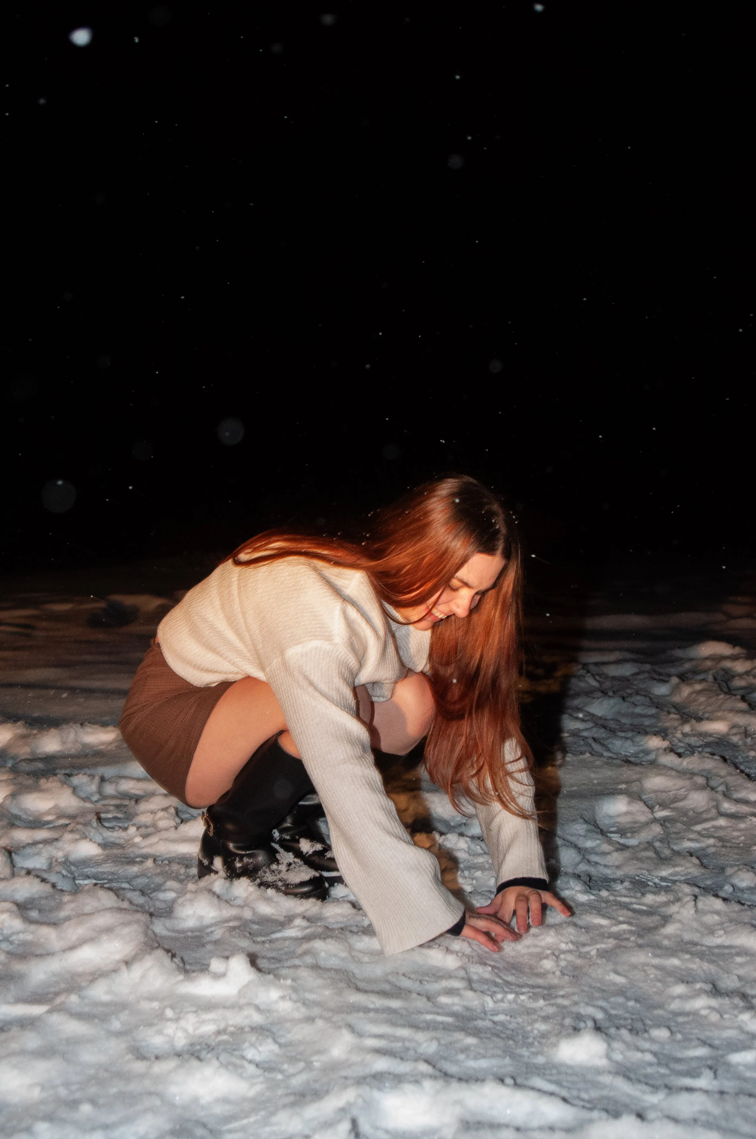 A woman with long red hair, wearing a beige sweater, brown skirt, and black boots, kneeling in the snow at night, looking down at the snow.