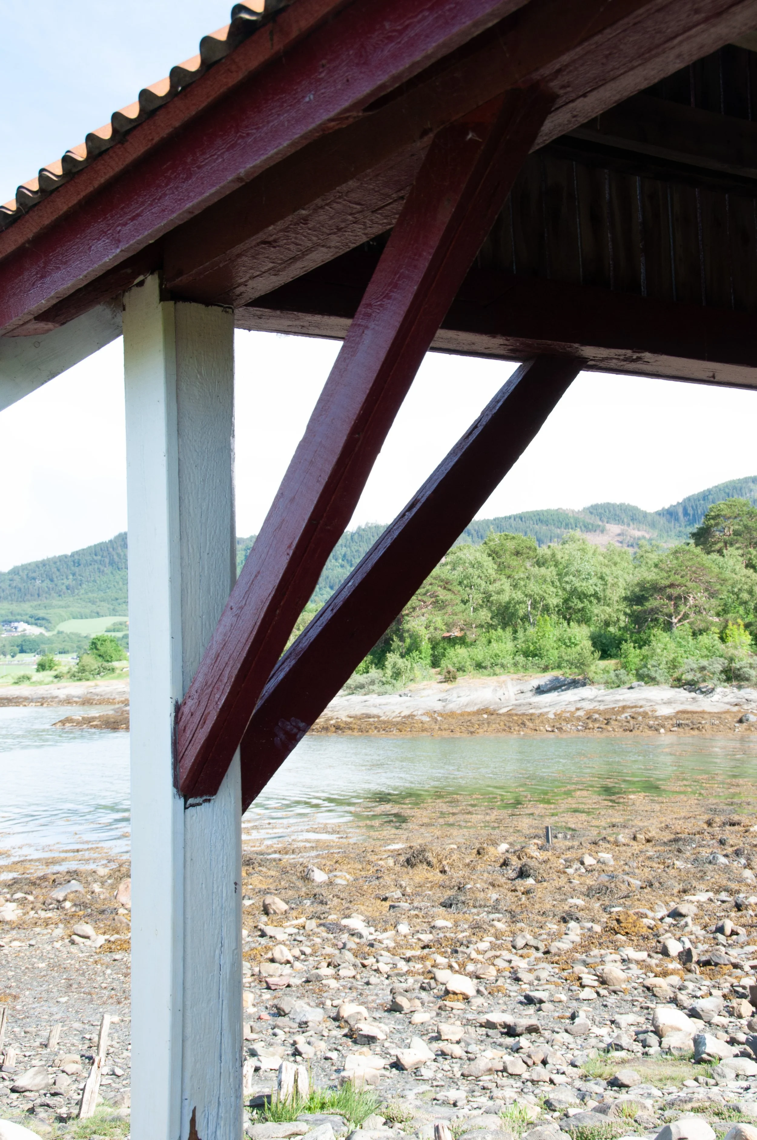 Close-up of a wooden pier or deck with white and red painted supports, overlooking a rocky shoreline with water, trees, and a mountain background.