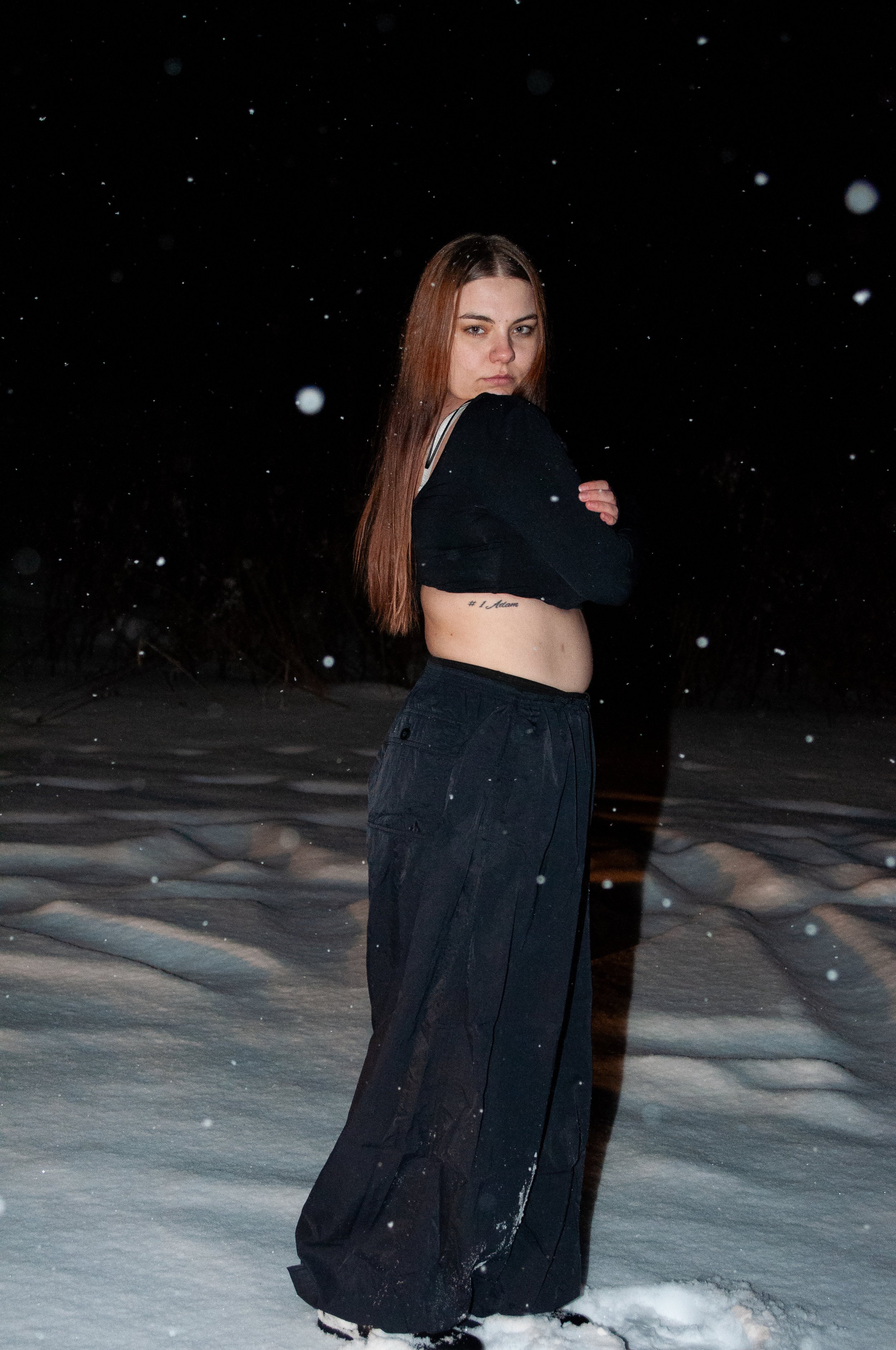 A woman stands outside on snow-covered ground at night, wearing black pants and a black jacket, with her arms crossed and looking at the camera.