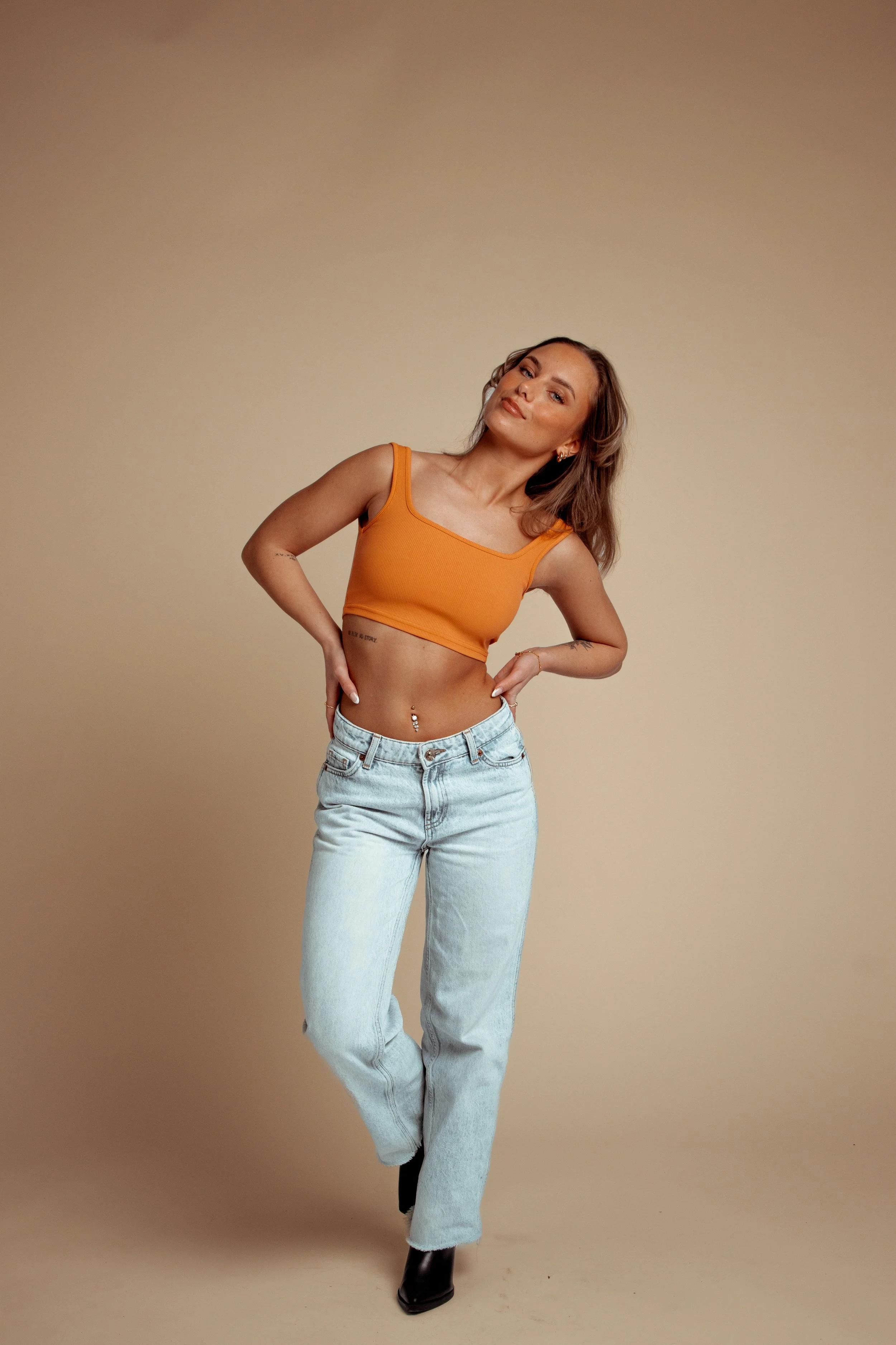 A woman wearing an orange crop top, light blue jeans, and black shoes, standing against a beige background, looking at the camera with a confident expression.