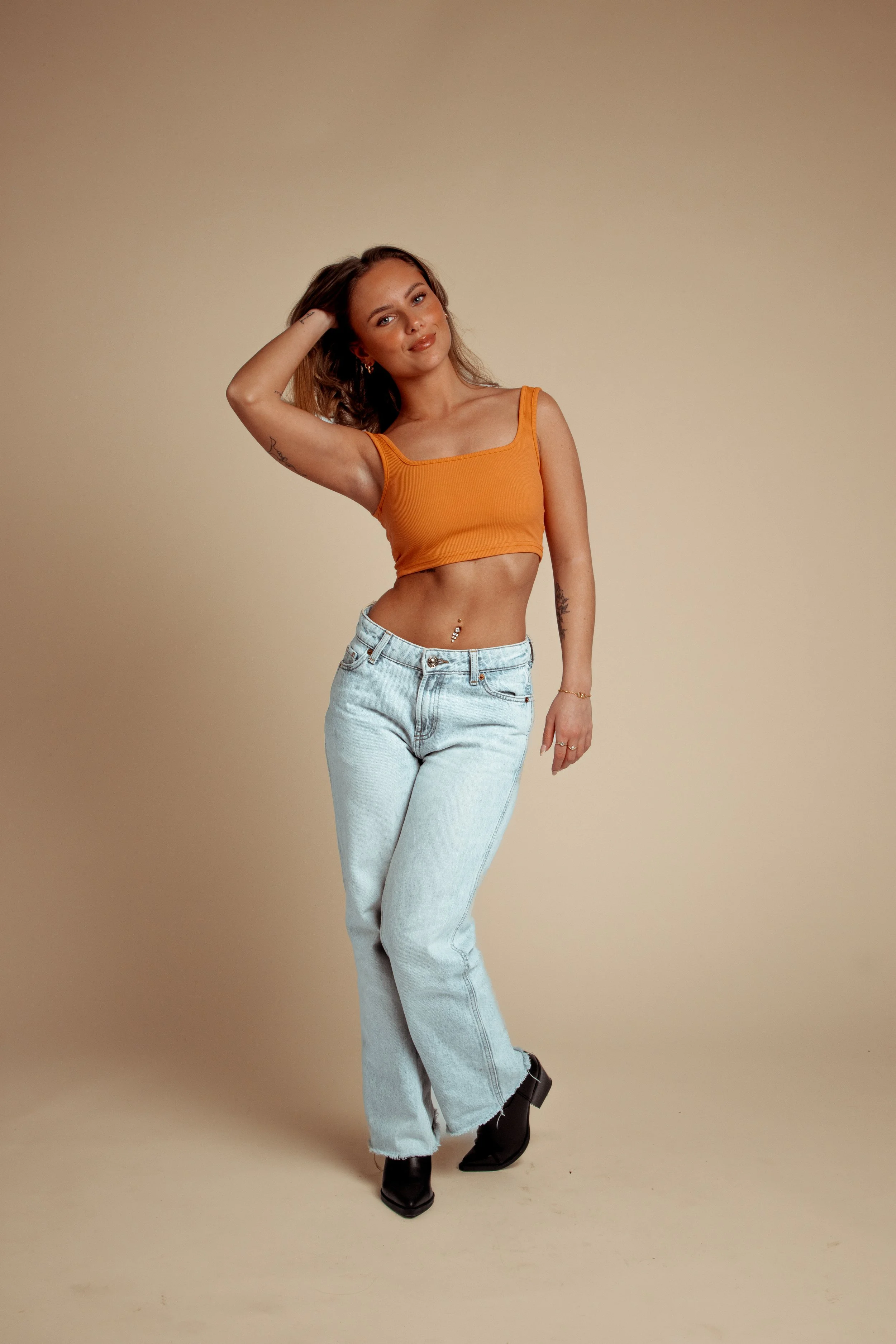 A young woman wearing a sleeveless orange crop top, light blue jeans, and black pointed-toe boots, standing against a plain beige background, posing with one hand in her hair and a slight smile.