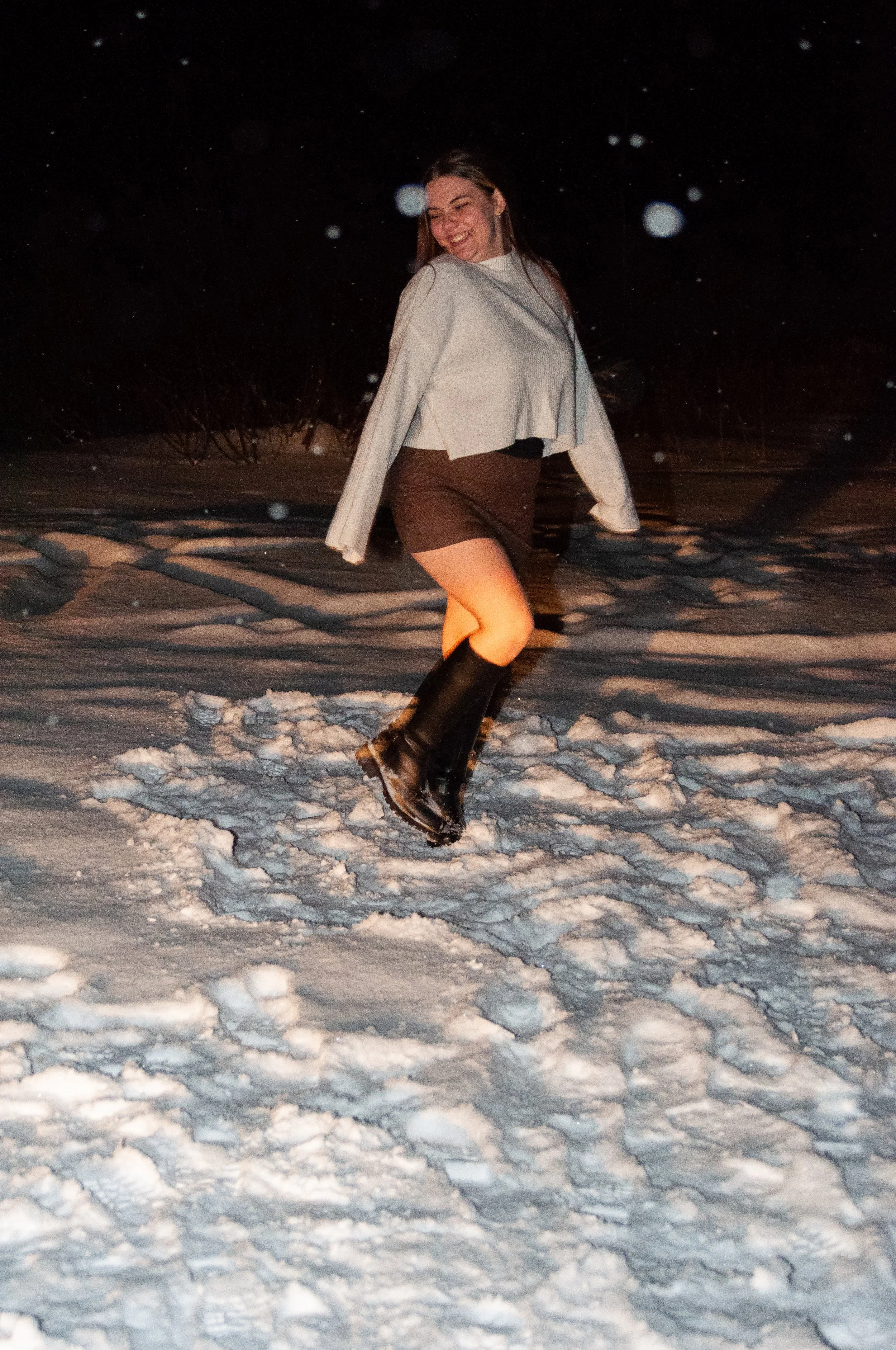 A woman in a white sweater, black skirt, and tall black boots dancing or playing in the snow at night.