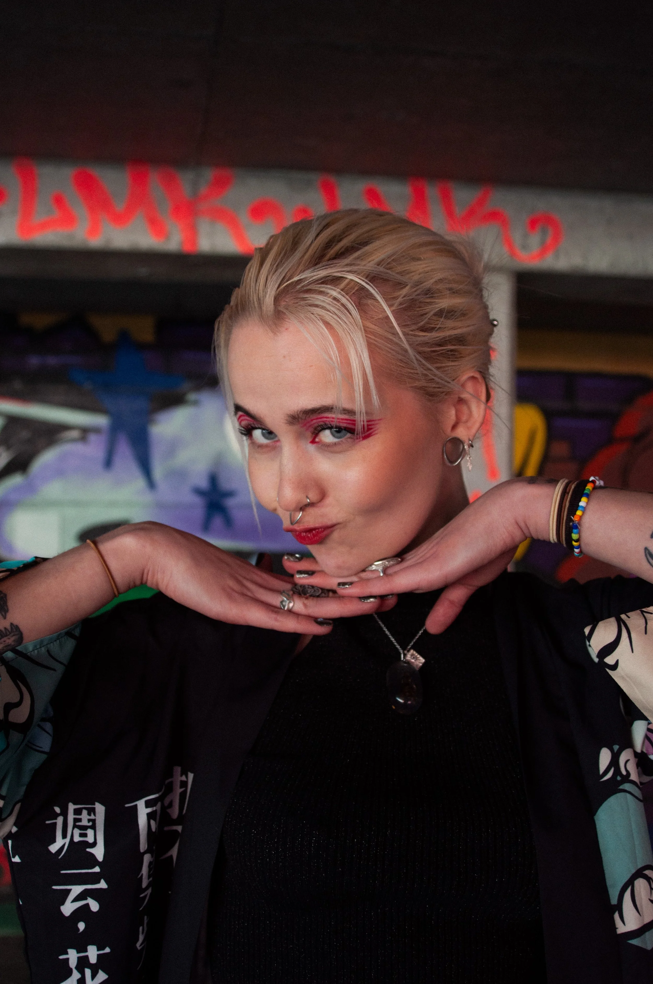 A young woman with blonde hair styled back, wearing vibrant makeup with pink eyeshadow and red lipstick, posing with her hands under her chin in front of a colorful, graffiti-covered background.