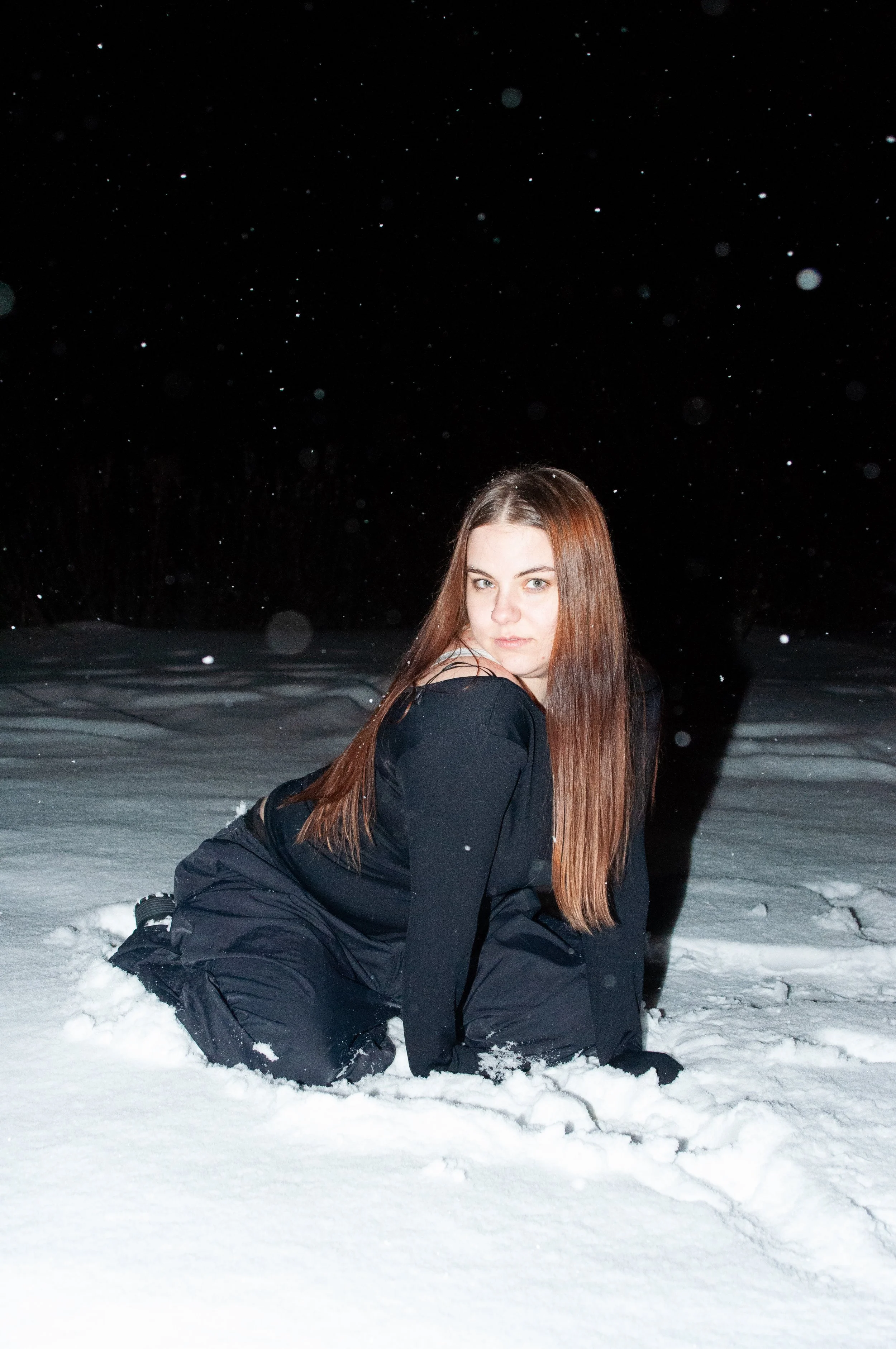 A young woman with long brown hair kneeling in the snow at night, wearing black outdoor clothing, with snow falling around her.