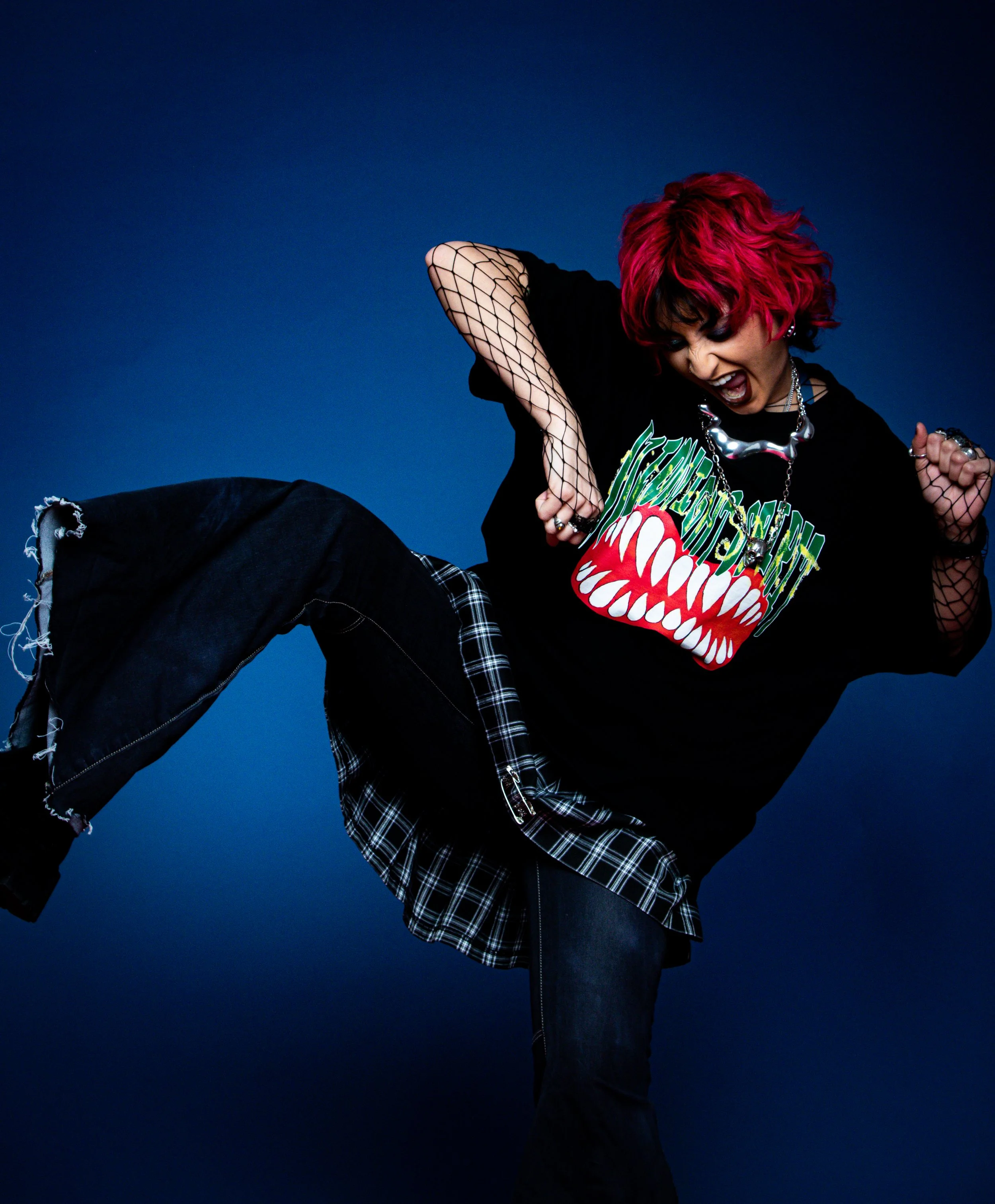 A woman with bright pink hair, wearing a black graphic t-shirt, fishnet sleeves, plaid skirt layered over ripped jeans, and jewelry, is striking a dynamic pose against a blue background.