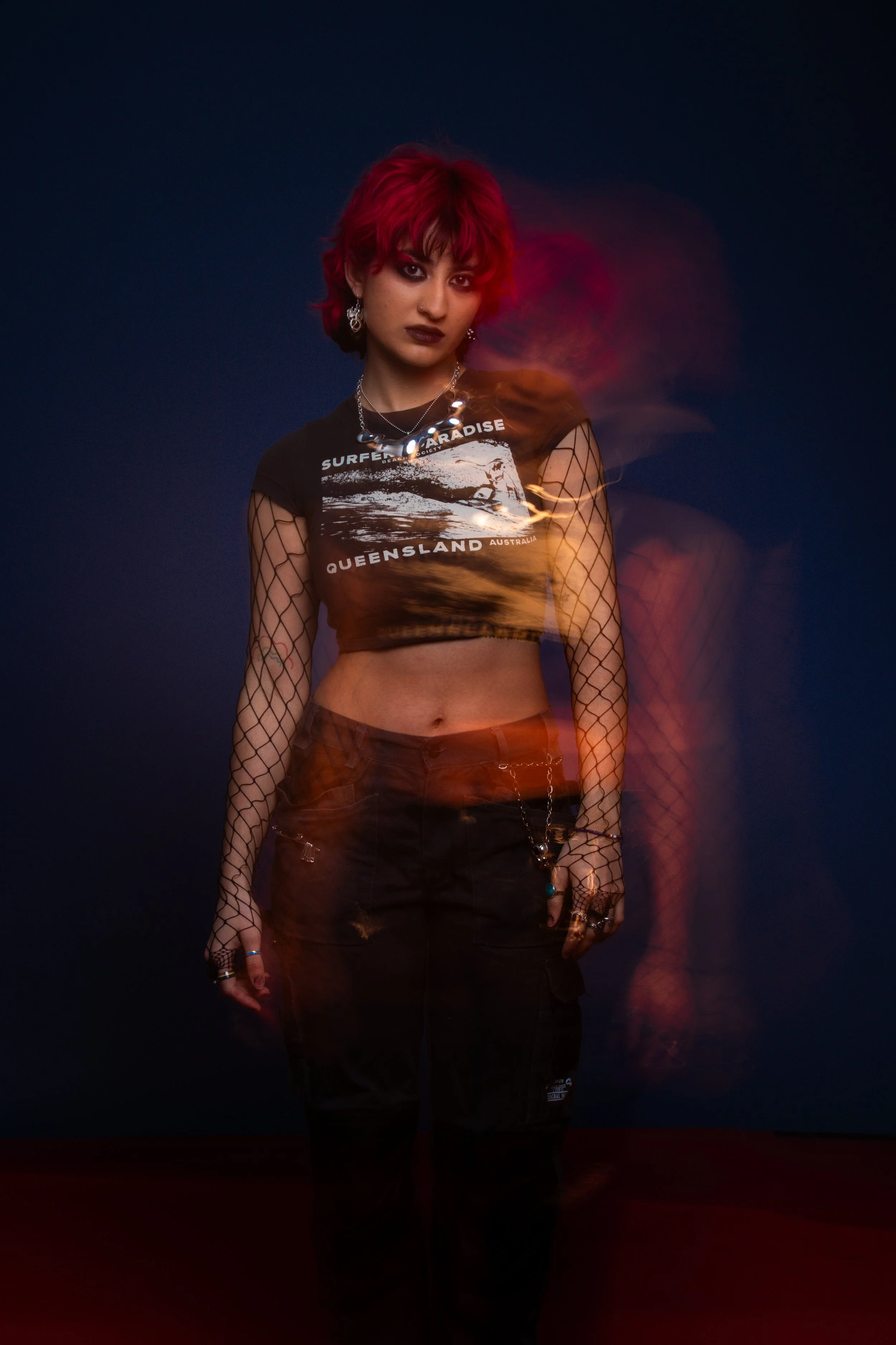 A young woman with short red hair and dark makeup posing against a dark background, wearing a black graphic crop top, fishnet sleeves, black pants, and jewelry.