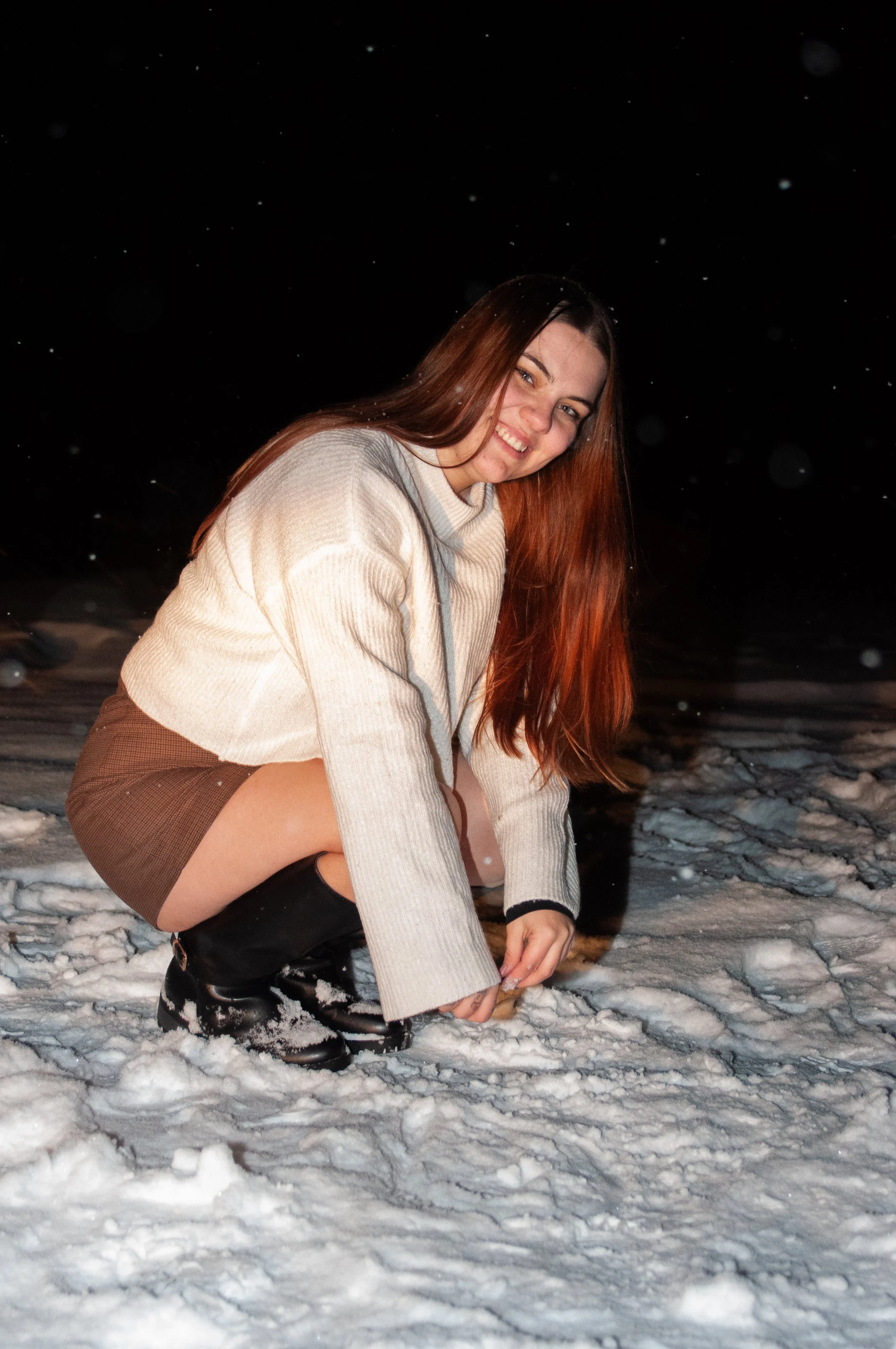 A woman with long brown hair squatting in the snow at night, smiling at the camera, wearing a beige sweater, a brown skirt, and black boots.