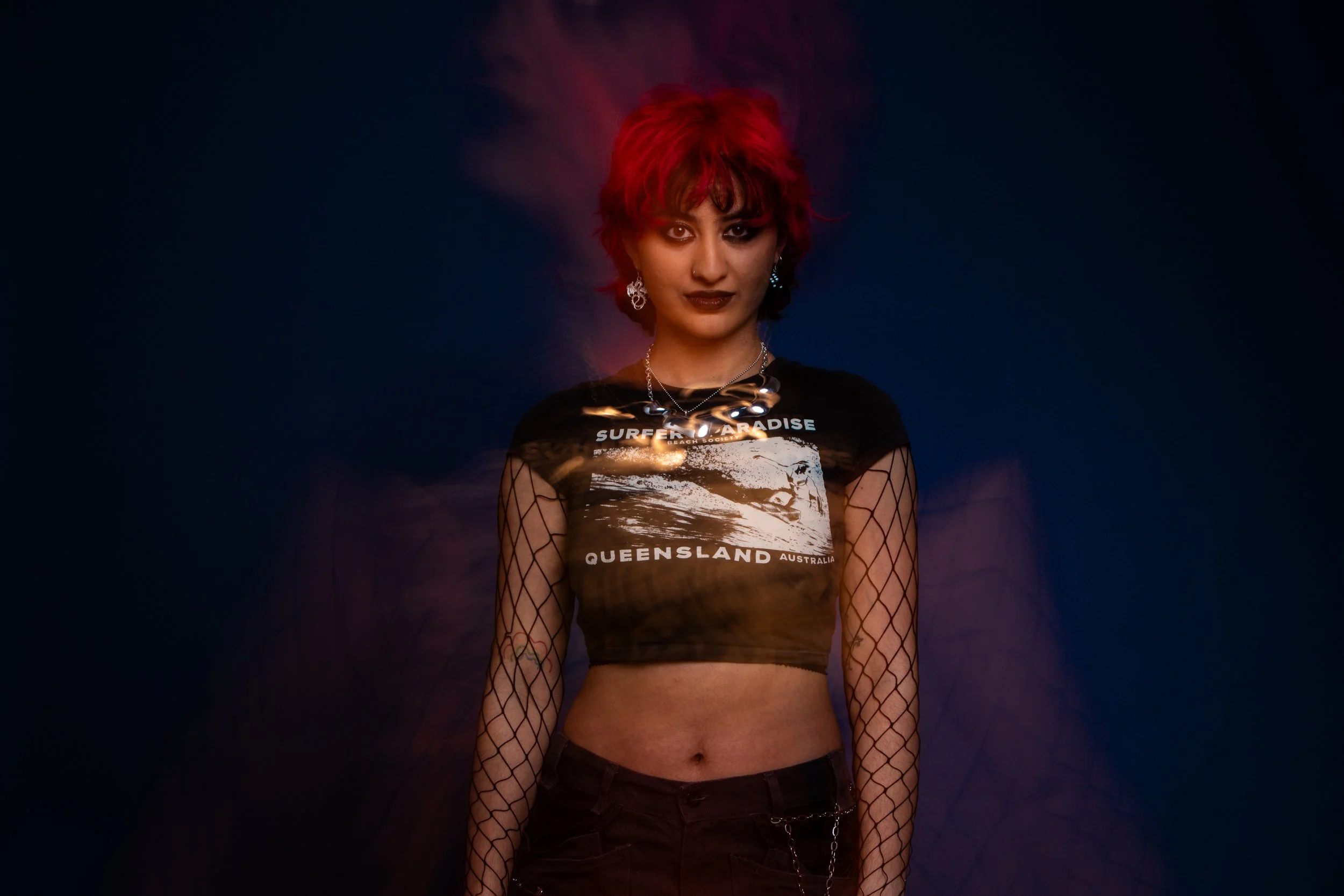 A woman with short red hair, dark makeup, and piercings, wearing a black mesh long-sleeve top with a graphic 'Surfer's Paradise' T-shirt underneath, posing against a dark background with purple and blue light effects.