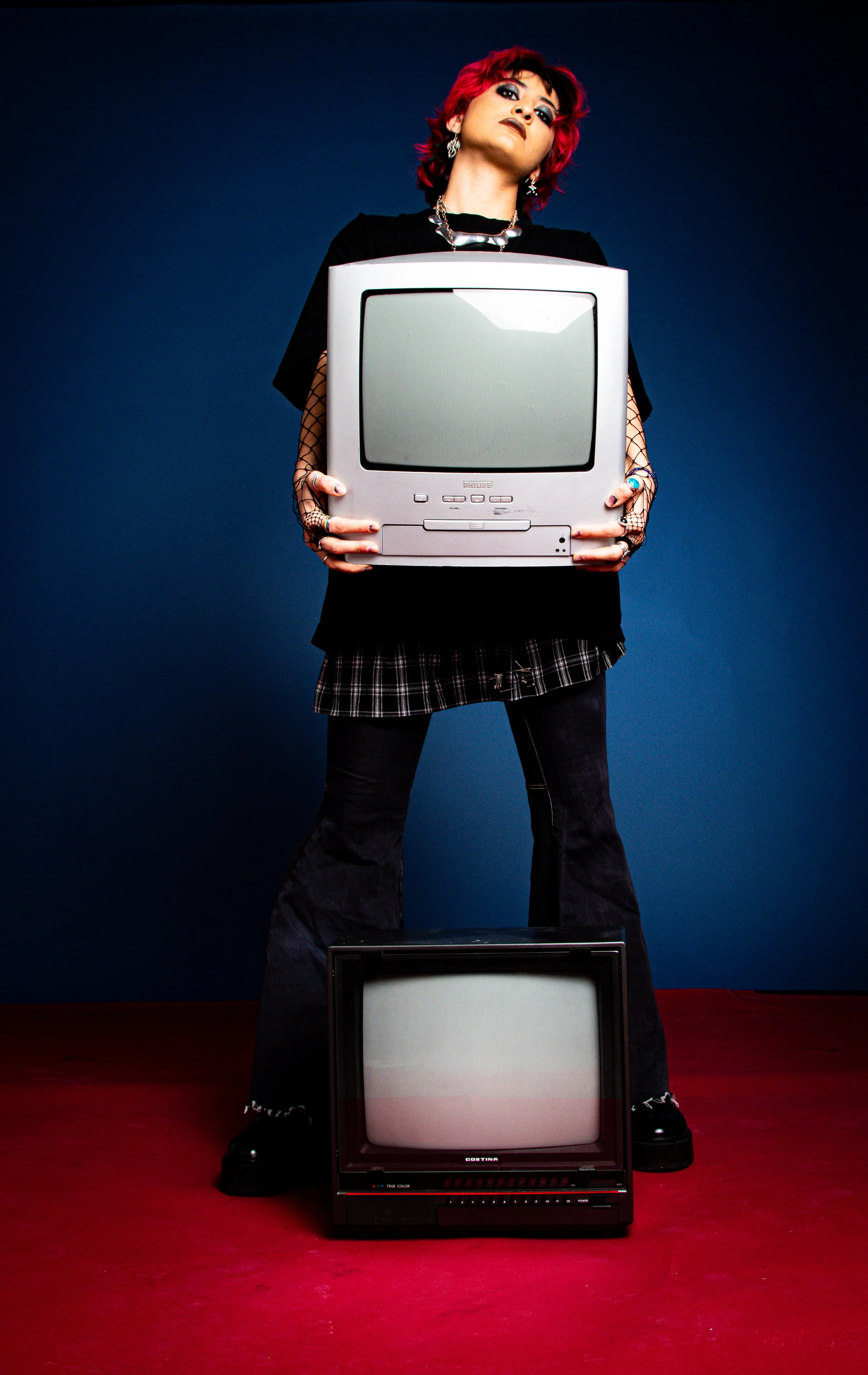 Person with bright red hair and punk style clothing holding an old CRT computer monitor, standing in front of a blue and red background.