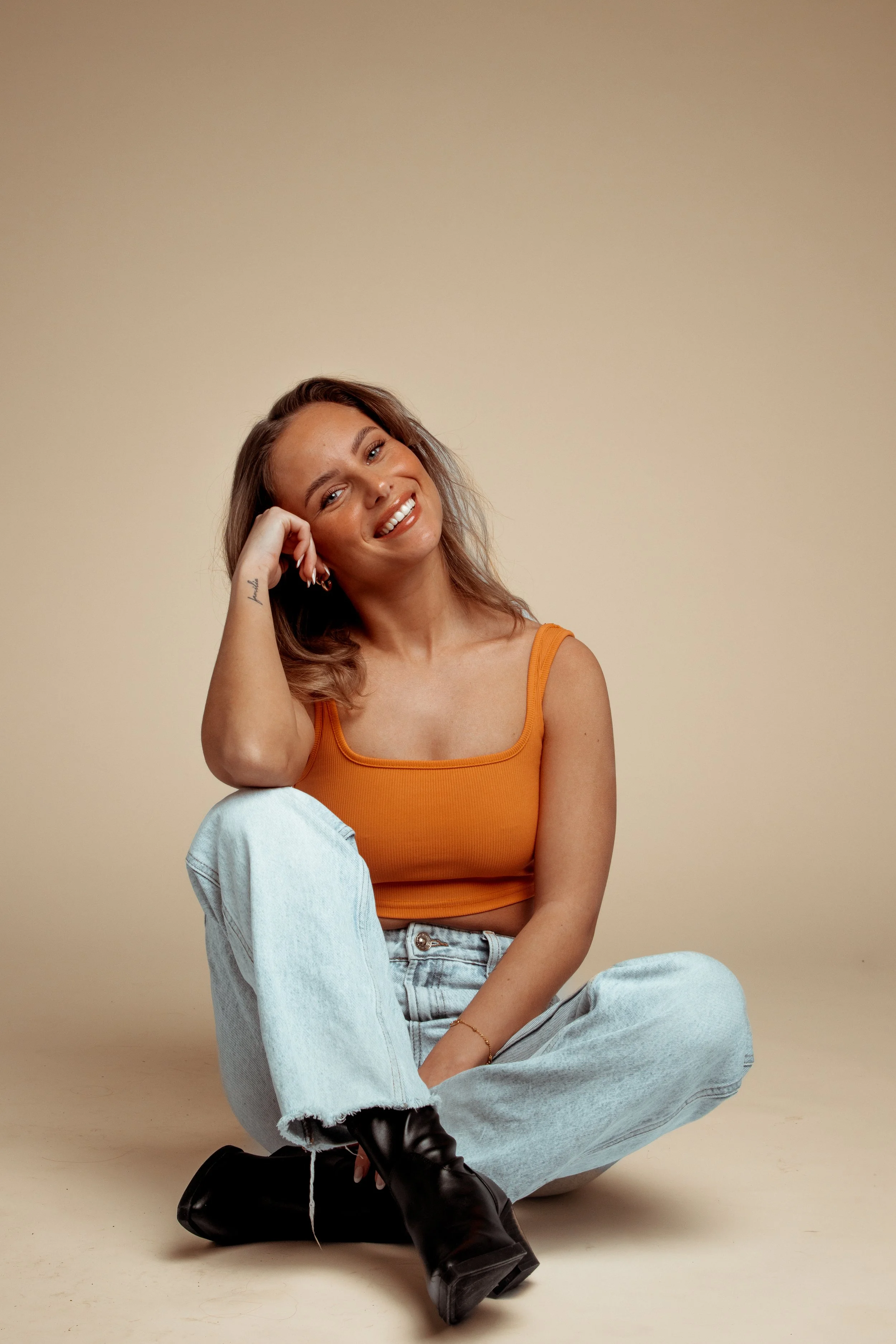 A young woman sitting on the floor, smiling, wearing an orange tank top, light blue jeans, and black boots, with a beige background.
