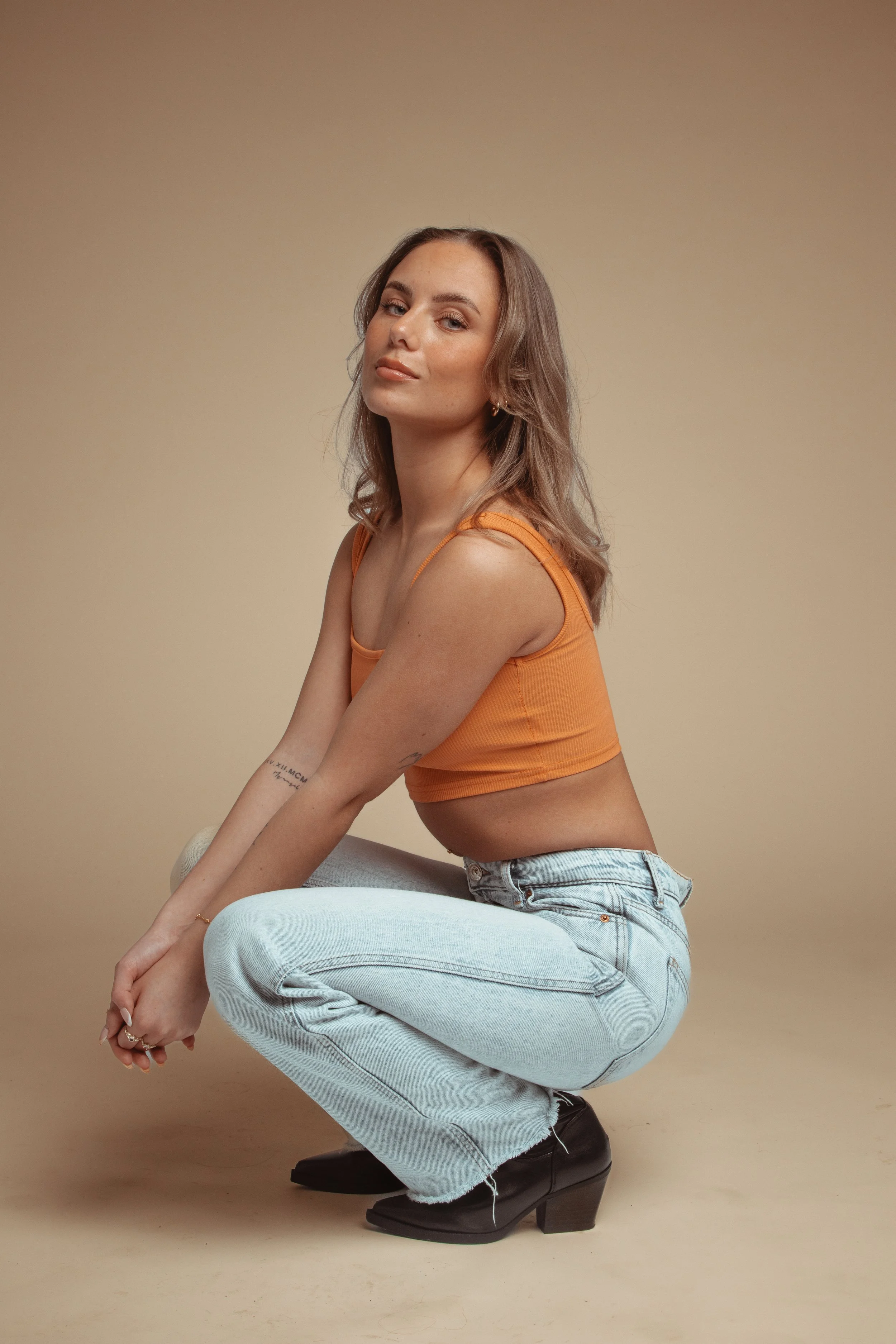 Young woman crouching in front of beige background, wearing an orange crop top, light blue jeans, and black heeled boots.