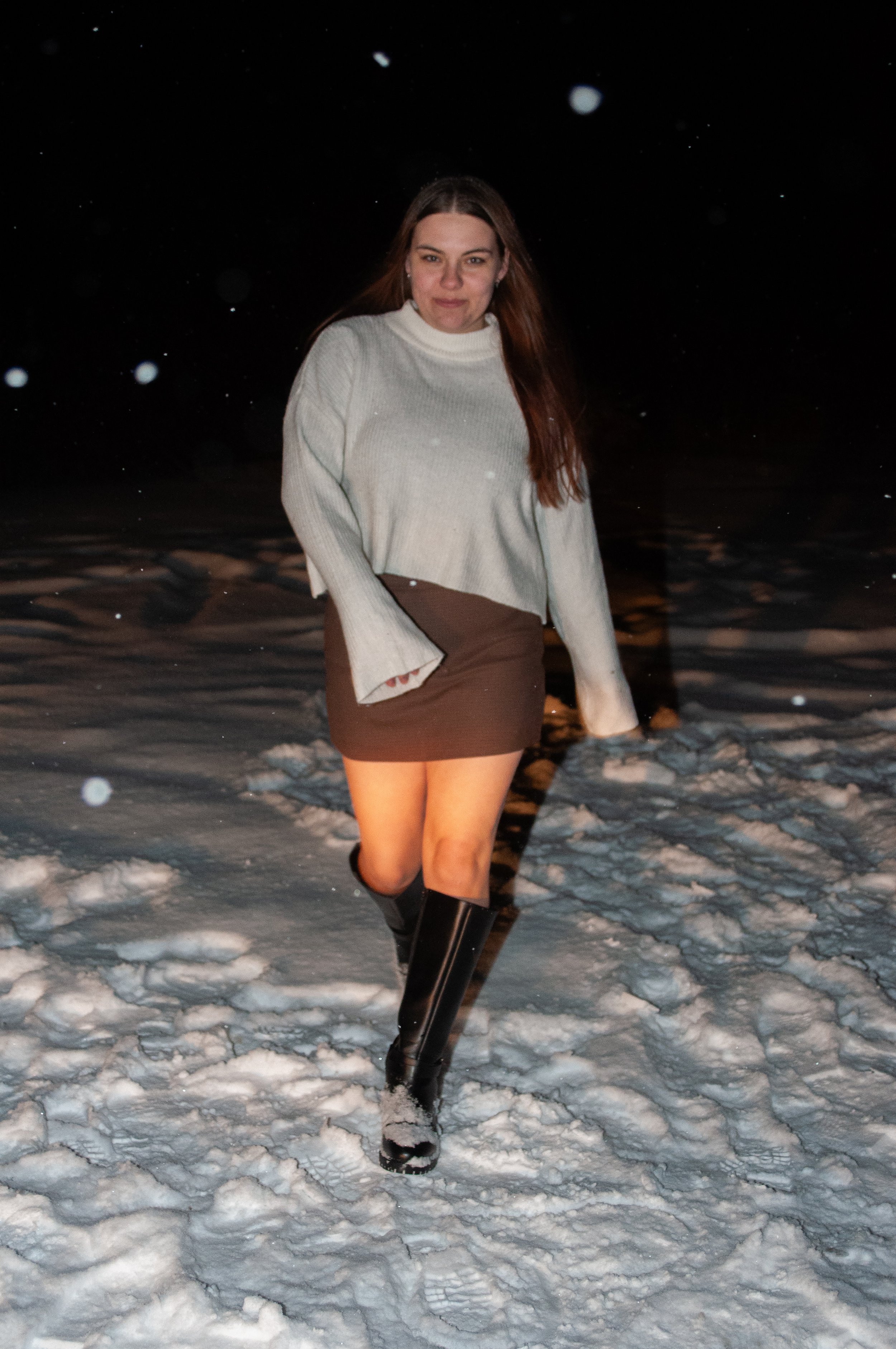 A woman in a white sweater, brown skirt, and knee-high black boots walking in snow at night.