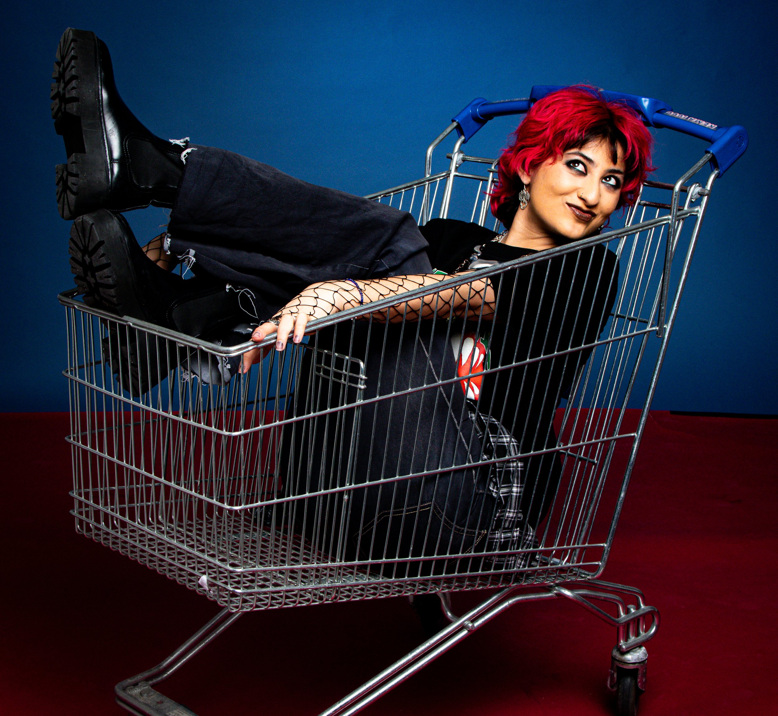 A woman with bright red hair, wearing black clothing including a fishnet sleeve, resting inside a shopping cart against a blue and red background.