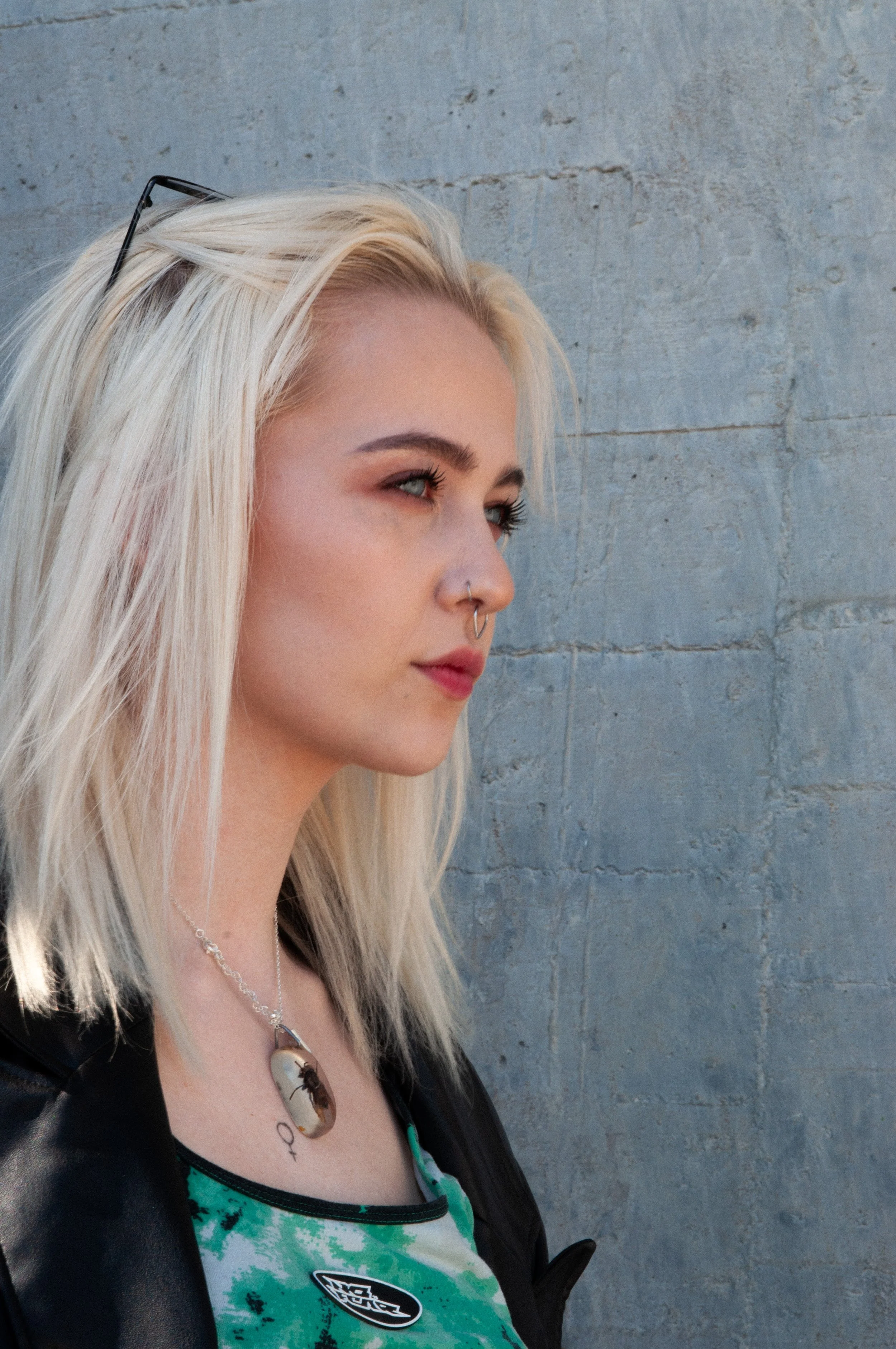A young woman with platinum blonde hair, wearing glasses on her head, a nose piercing, a necklace, and a green tie-dye shirt, standing against a textured concrete wall.