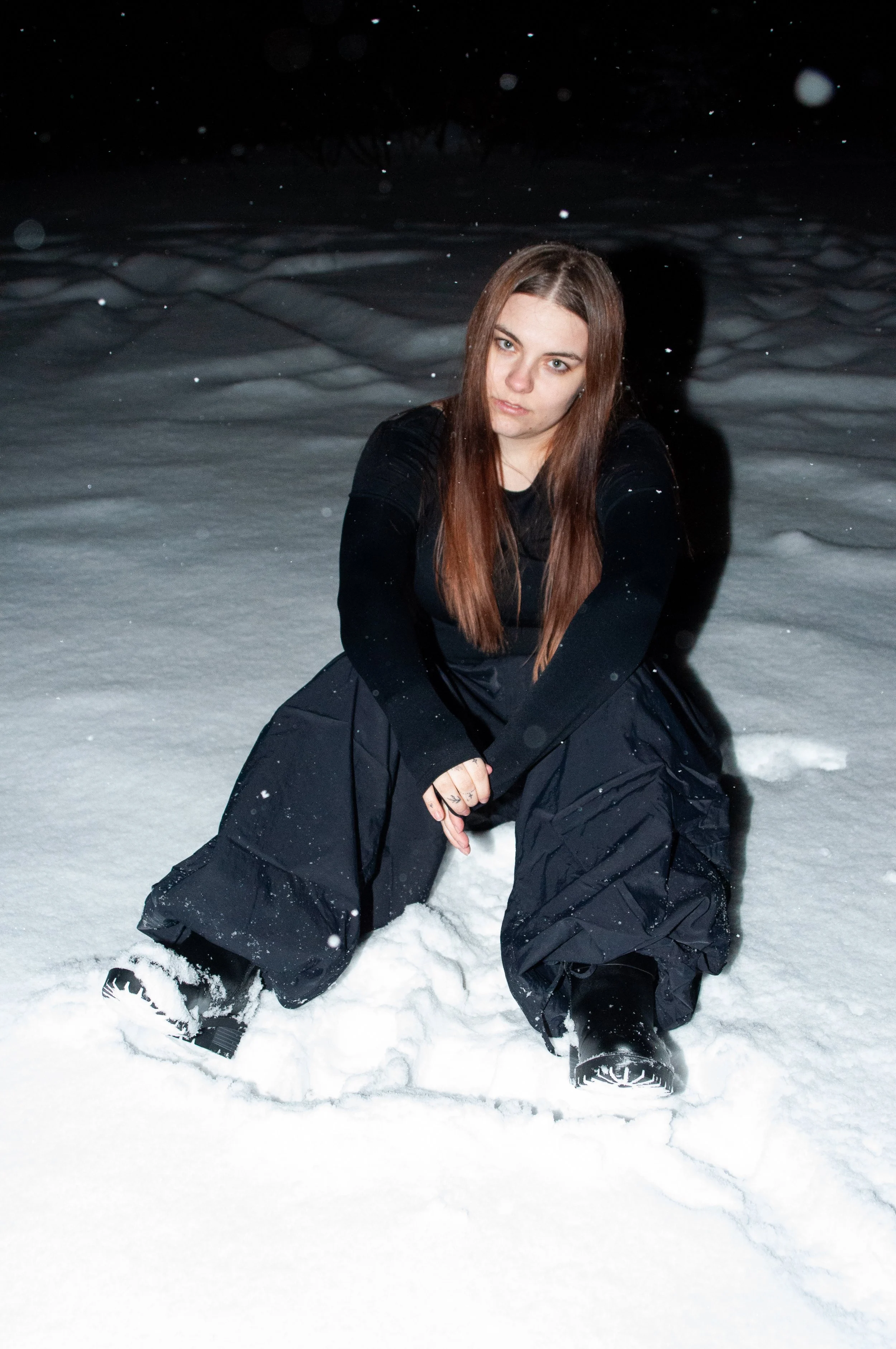A woman sitting in snow at night, wearing black clothing, with long brown hair and serious expression.