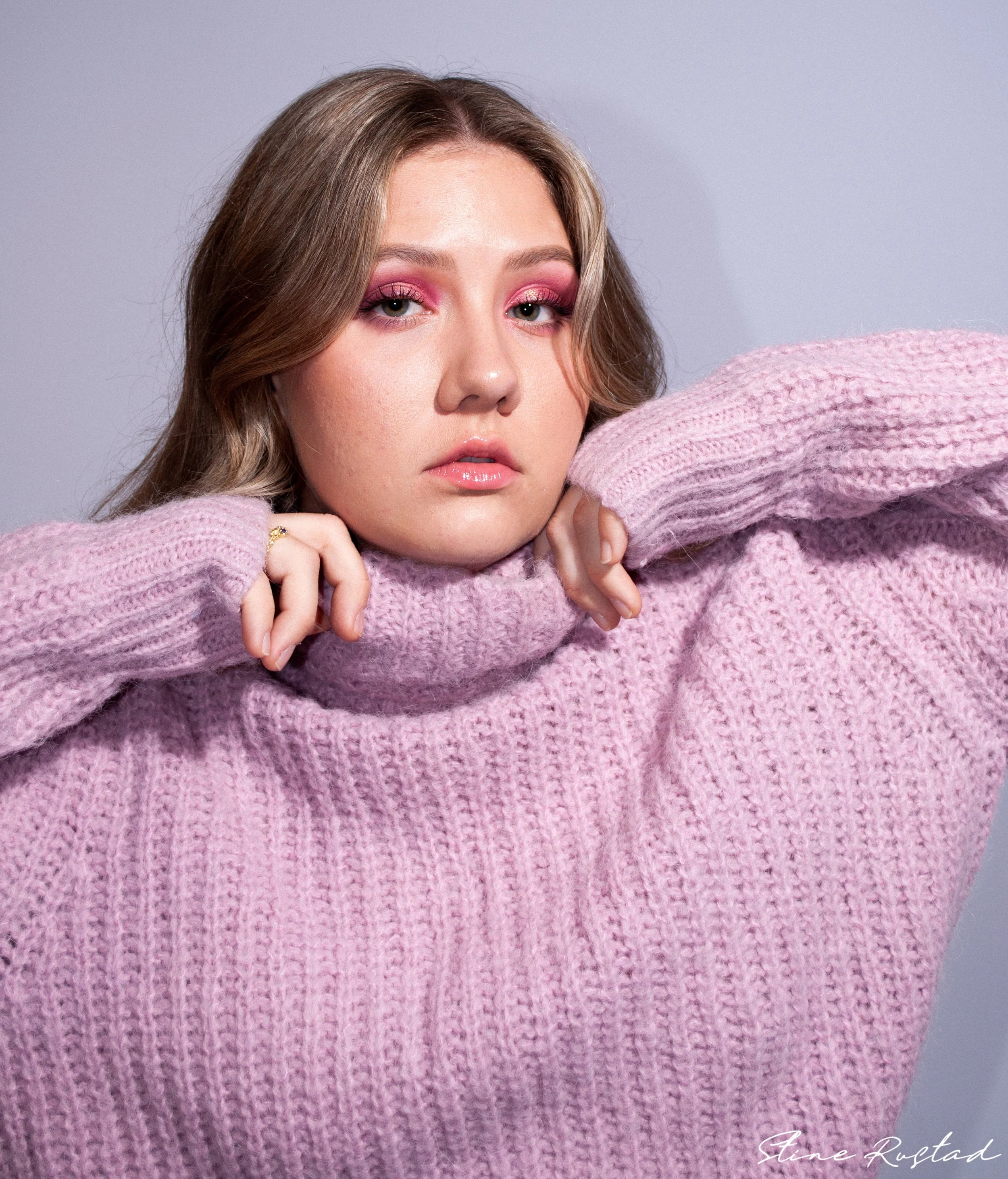 A young woman with light brown hair and pink eyeshadow wearing a pink knit sweater poses with her arms resting on a surface, looking into the camera with a neutral expression.