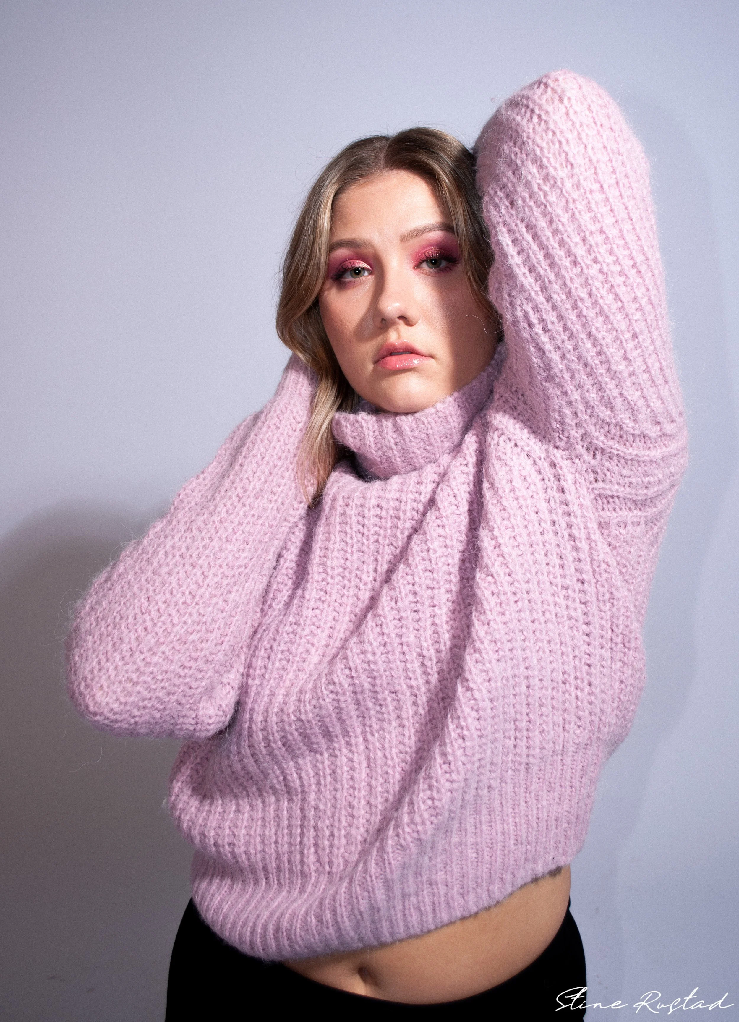 A young woman with wavy blonde hair is wearing a pink knit sweater and is posing with both arms raised above her head, one hand touching her hair and the other holding her neck. The background is plain and light-colored.