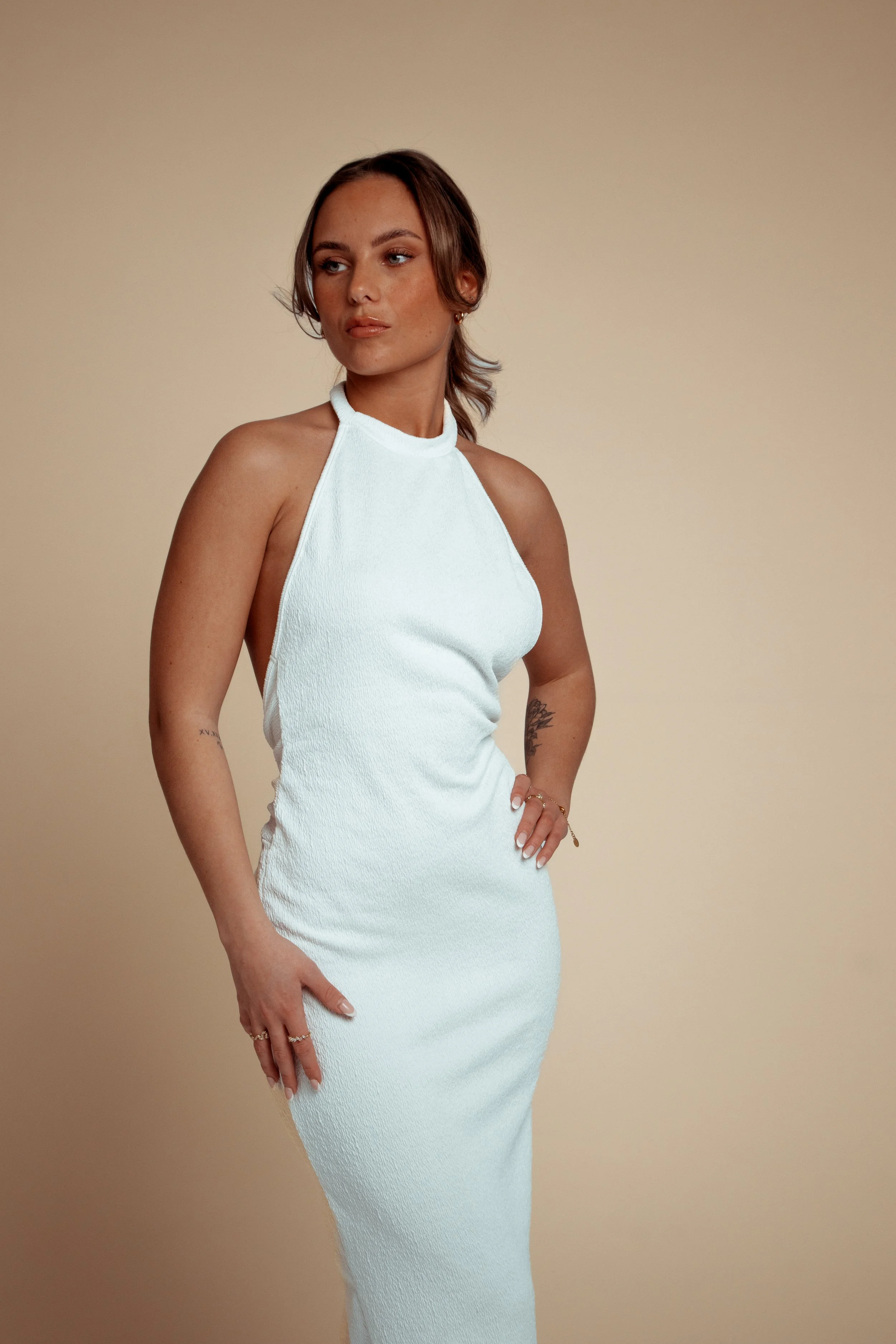 A woman wearing a sleeveless white dress with a high neck, standing against a plain beige background.